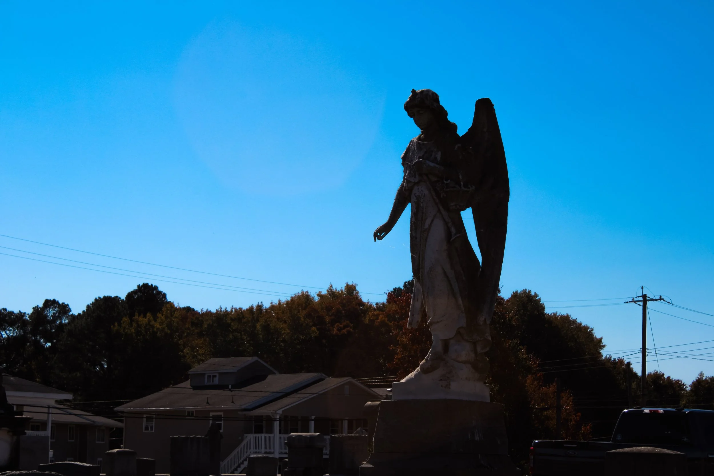 A statue of a woman with wings on her back, standing outdoors against a blue sky, with trees and houses in the background.