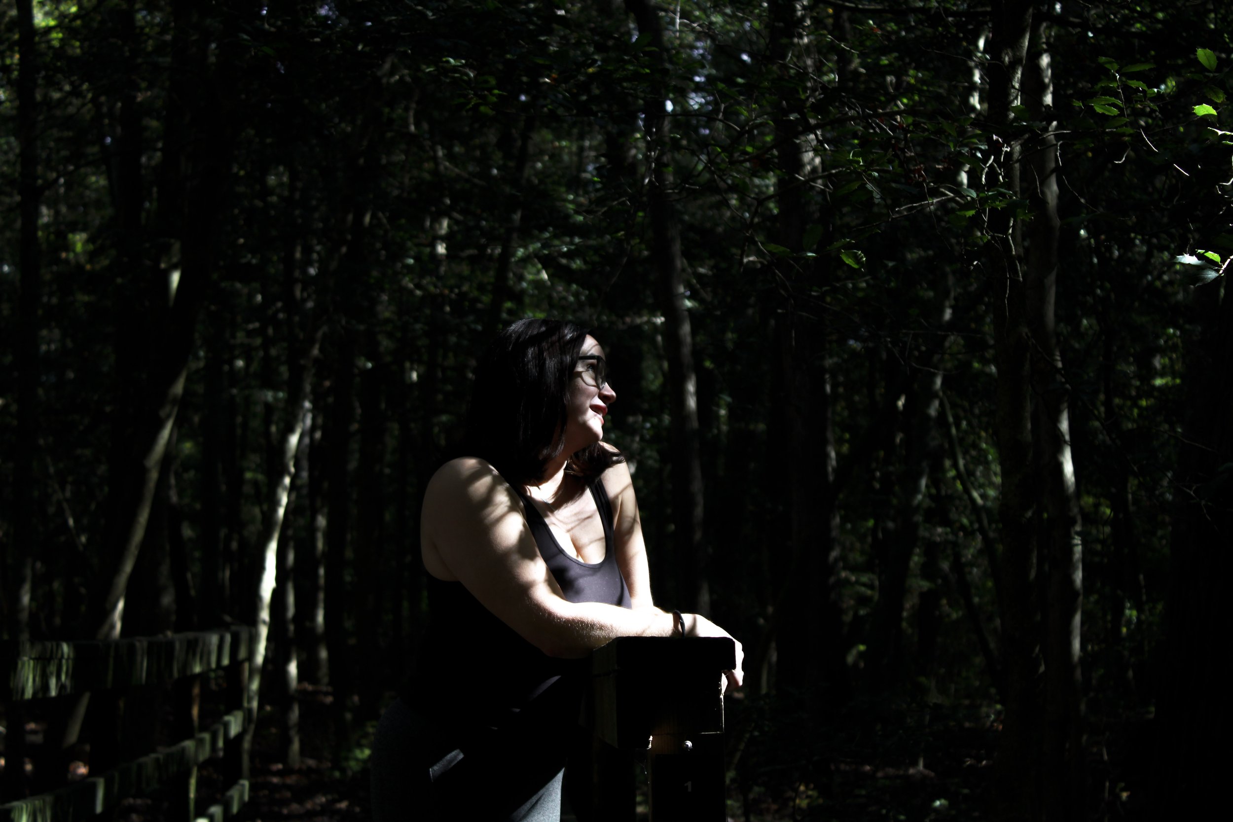 A woman with dark hair, glasses, and a black sleeveless top stands outdoors in a forested area with dense trees and green leaves, illuminated by sunlight.