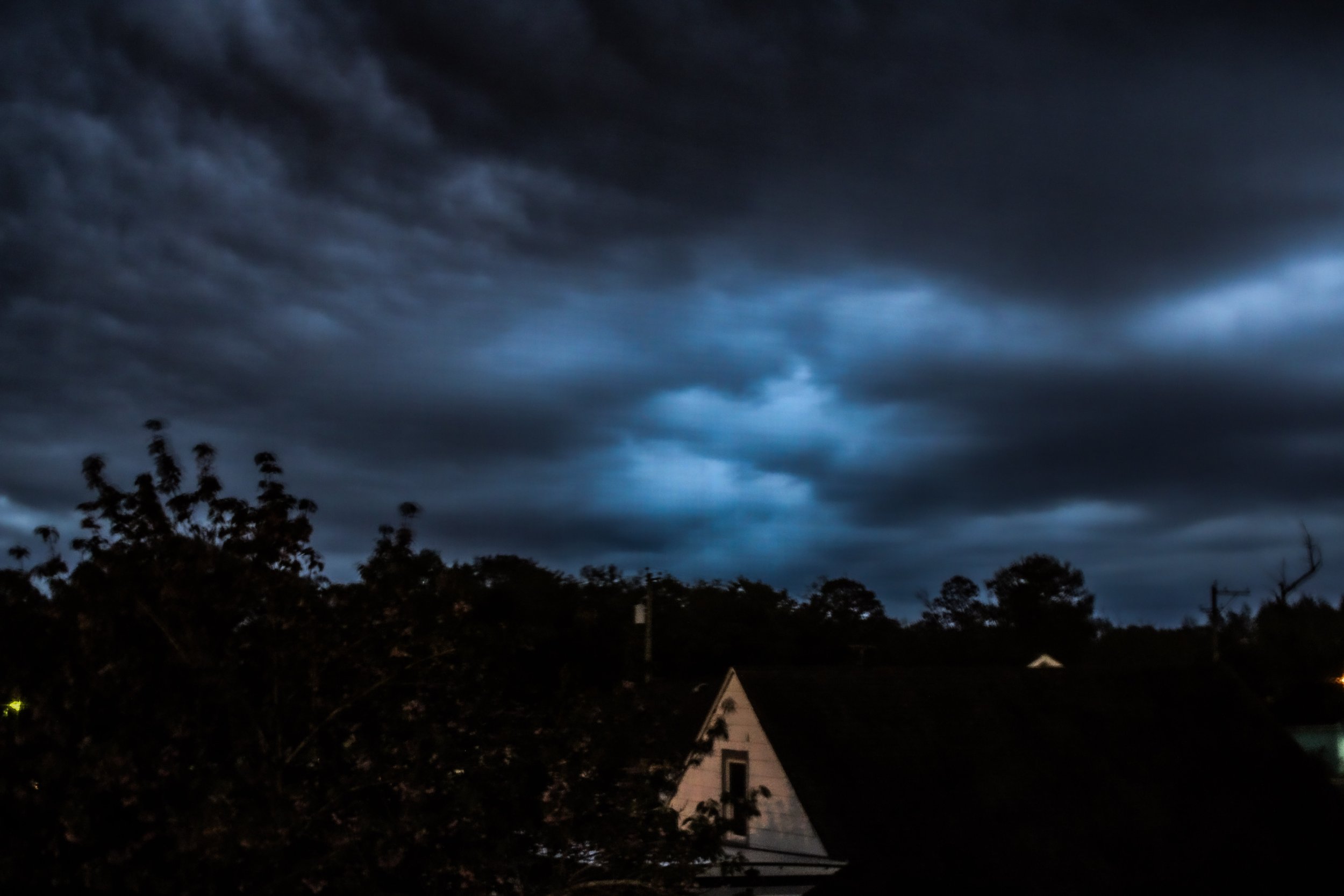 Dark, stormy clouds fill the sky at night, with silhouettes of trees and part of a house visible in the foreground.