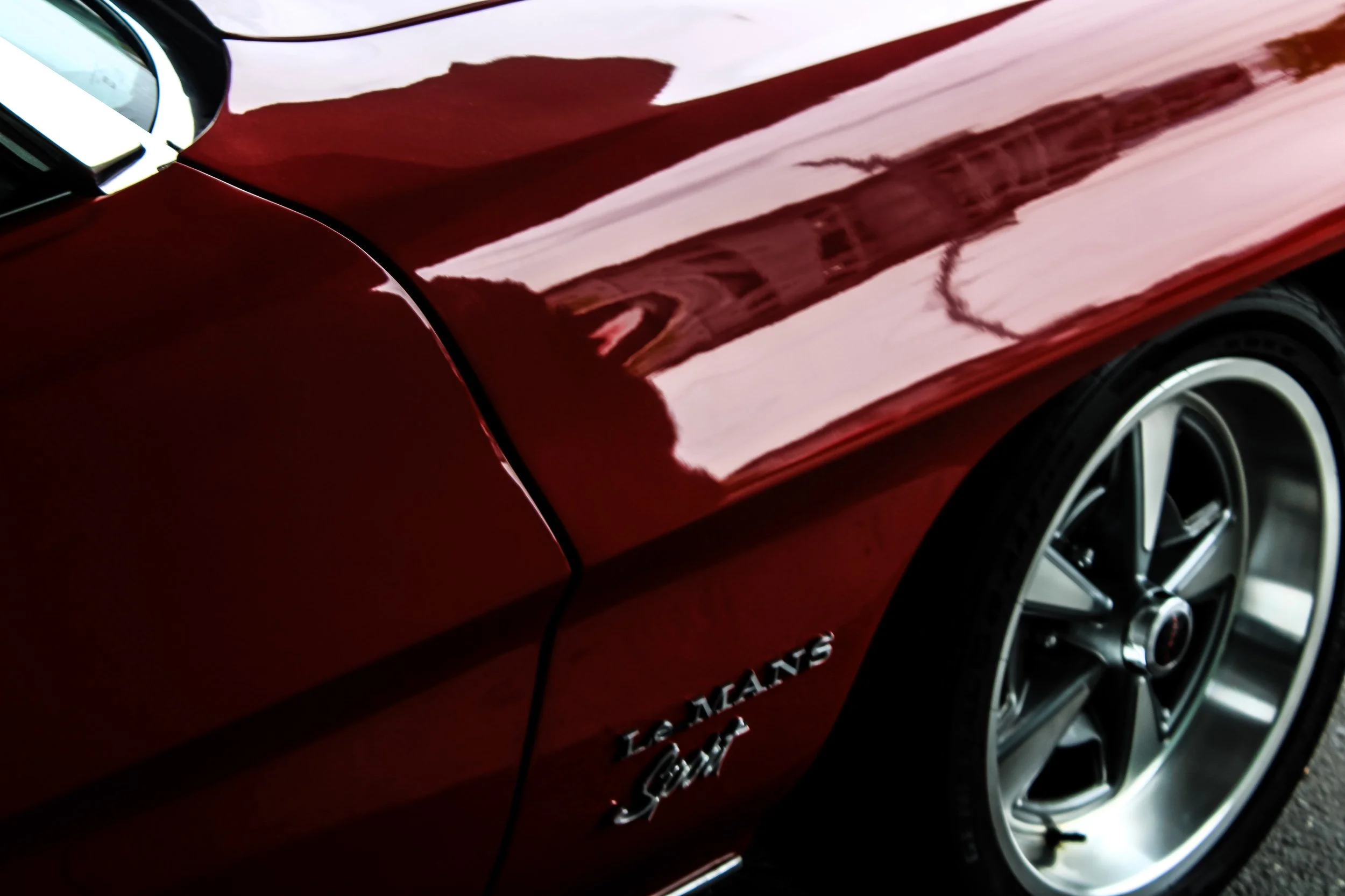 Close-up of a red car's side, showing reflections on the shiny surface and a wheel with a silver alloy rim.