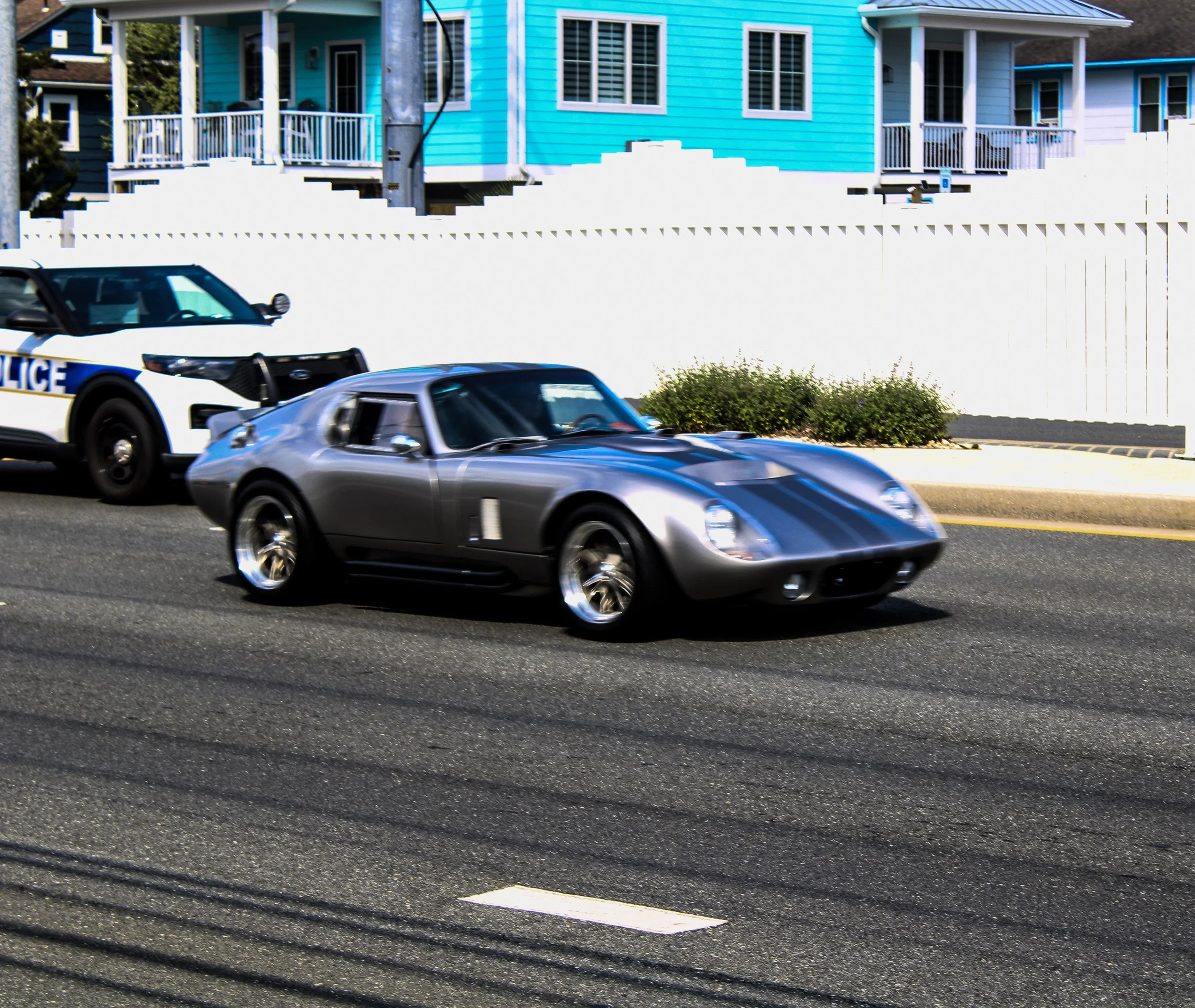 Street view with a silver sports car, police car, and a blue house with white trim in the background.