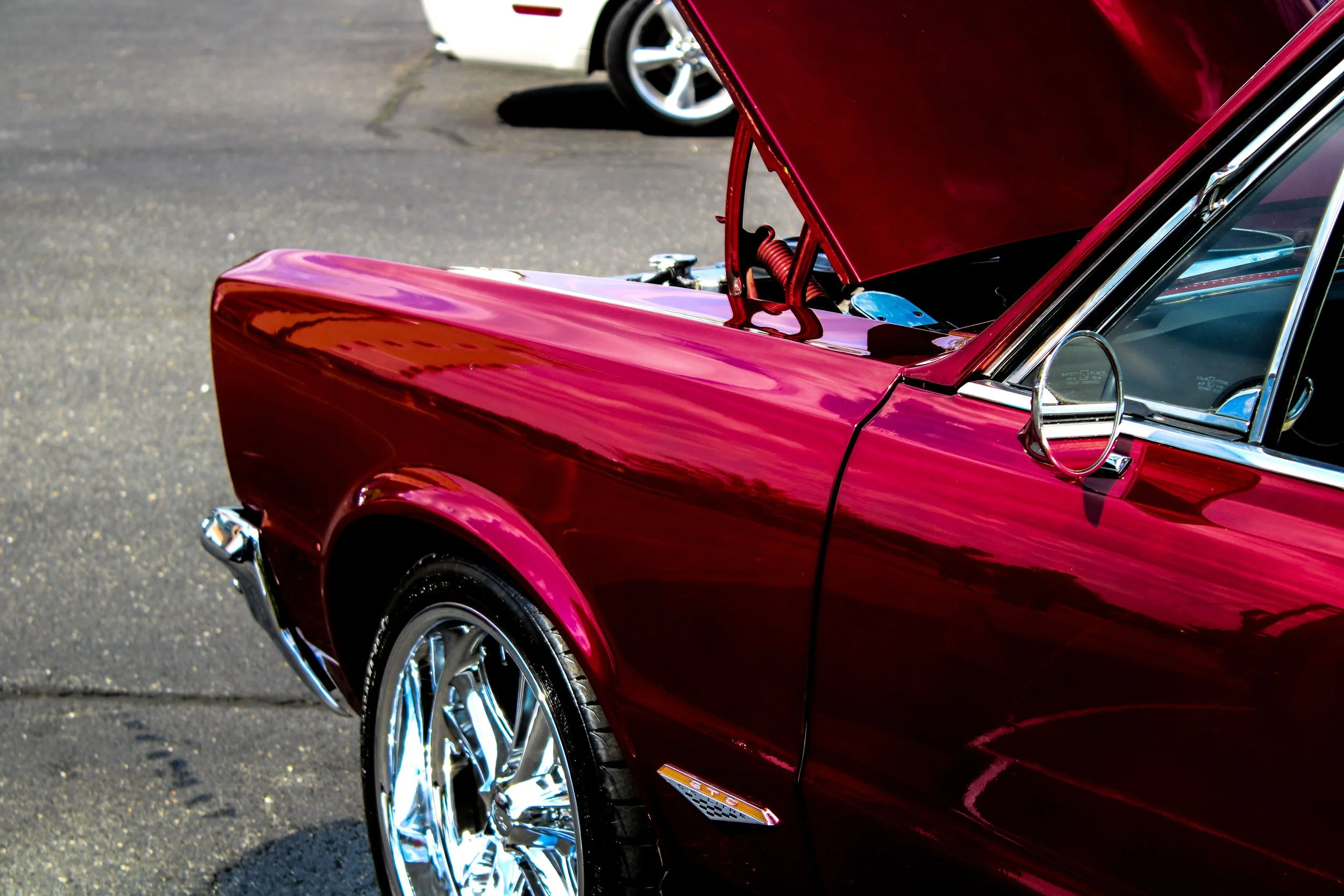 Close-up of a shiny red vintage car with open hood, parked on pavement, with another vehicle in the background.