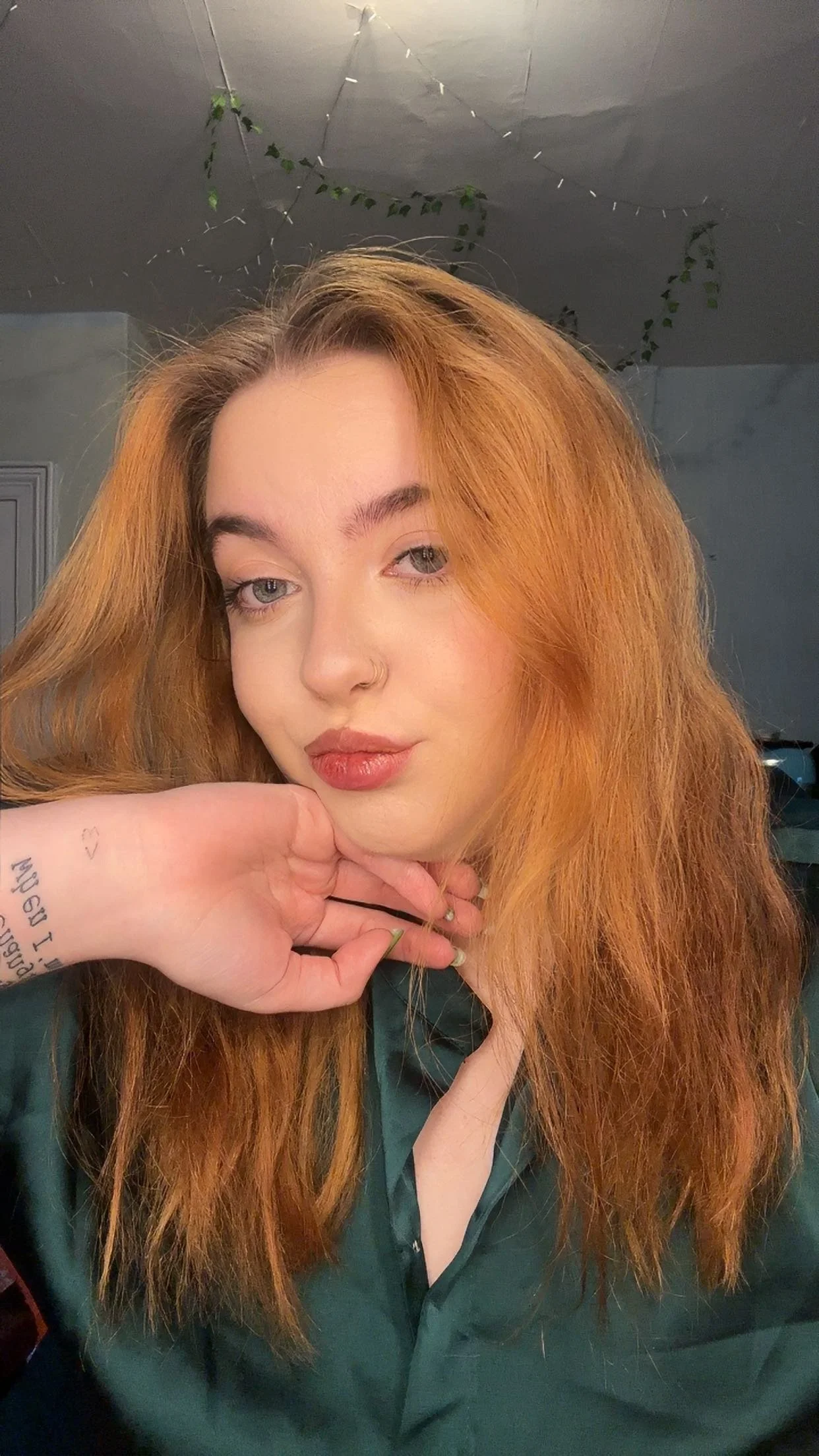 Close-up of a woman with long, wavy red hair, wearing a dark green top, resting her chin on her hand, looking at the camera. The background shows a ceiling with hanging plants.