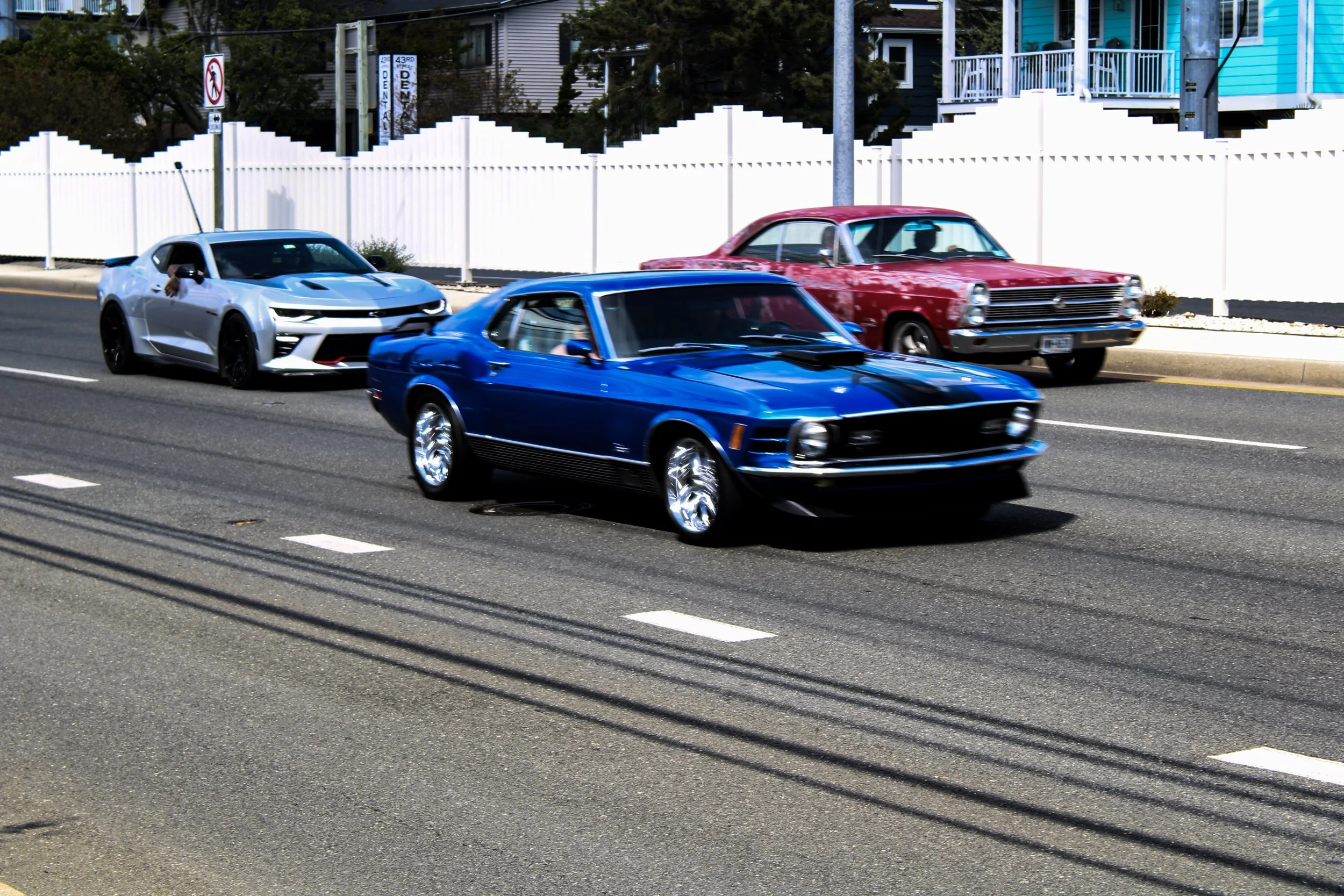 Three cars driving on a city street with a white picket fence and colorful houses in the background, one silver, one blue, and one red.