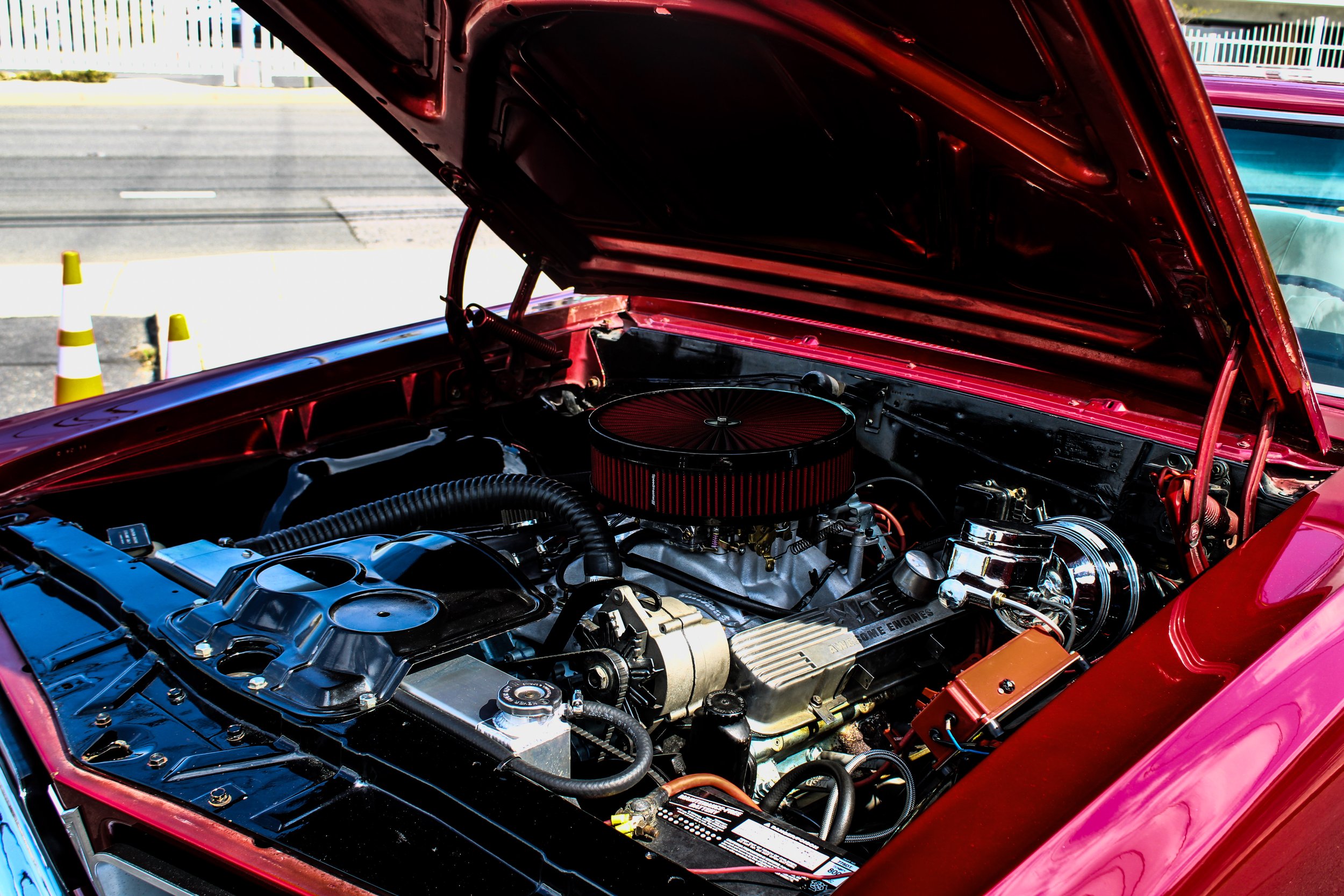 Red classic car with its hood open, displaying the engine bay and engine components, parked outdoors with a white fence and traffic cones in the background.