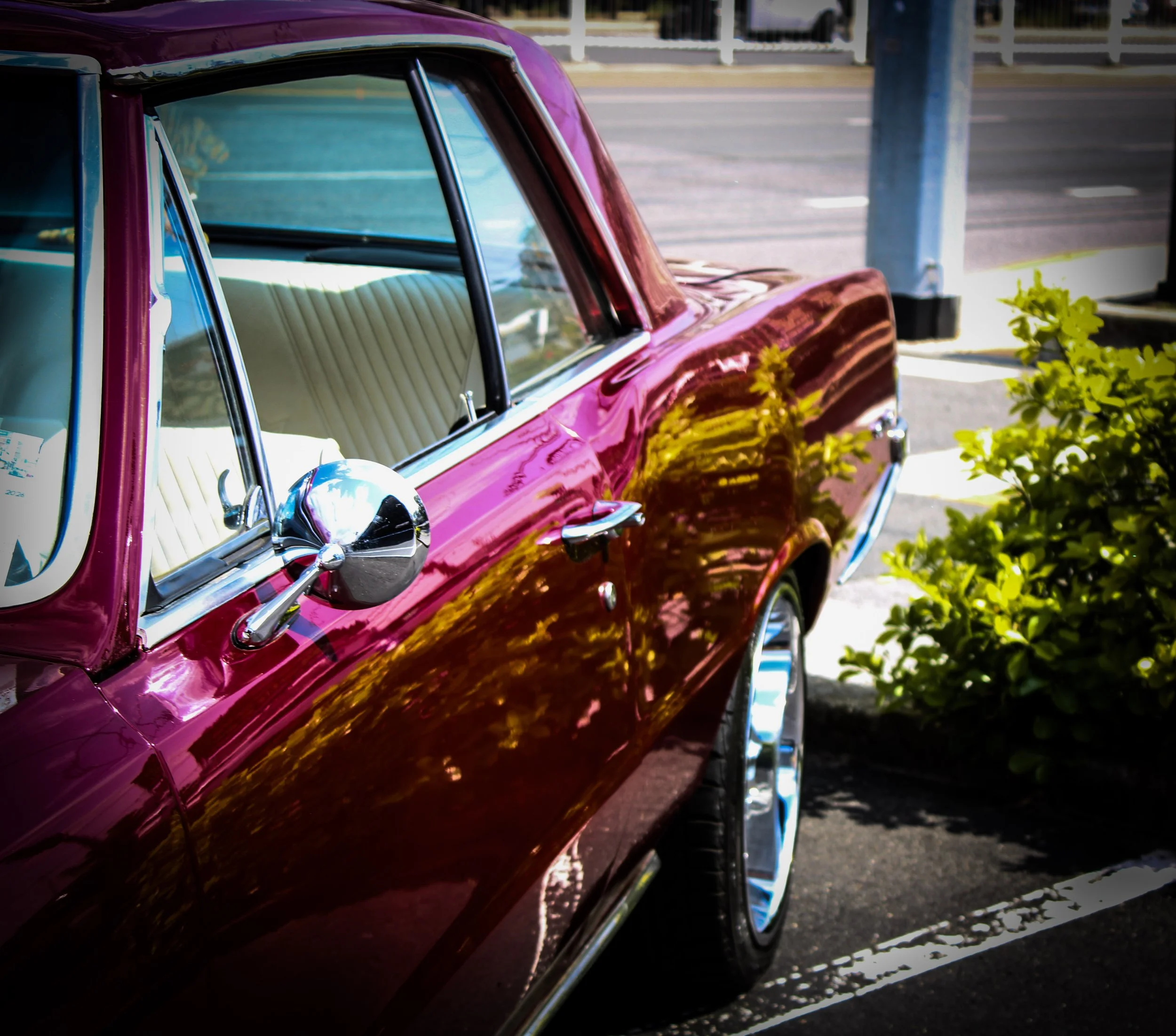 Close-up of a vintage pink car parked on the side of the street, with visible chrome side mirror and window details, in a sunny setting with some greenery and a parking lot in the background.