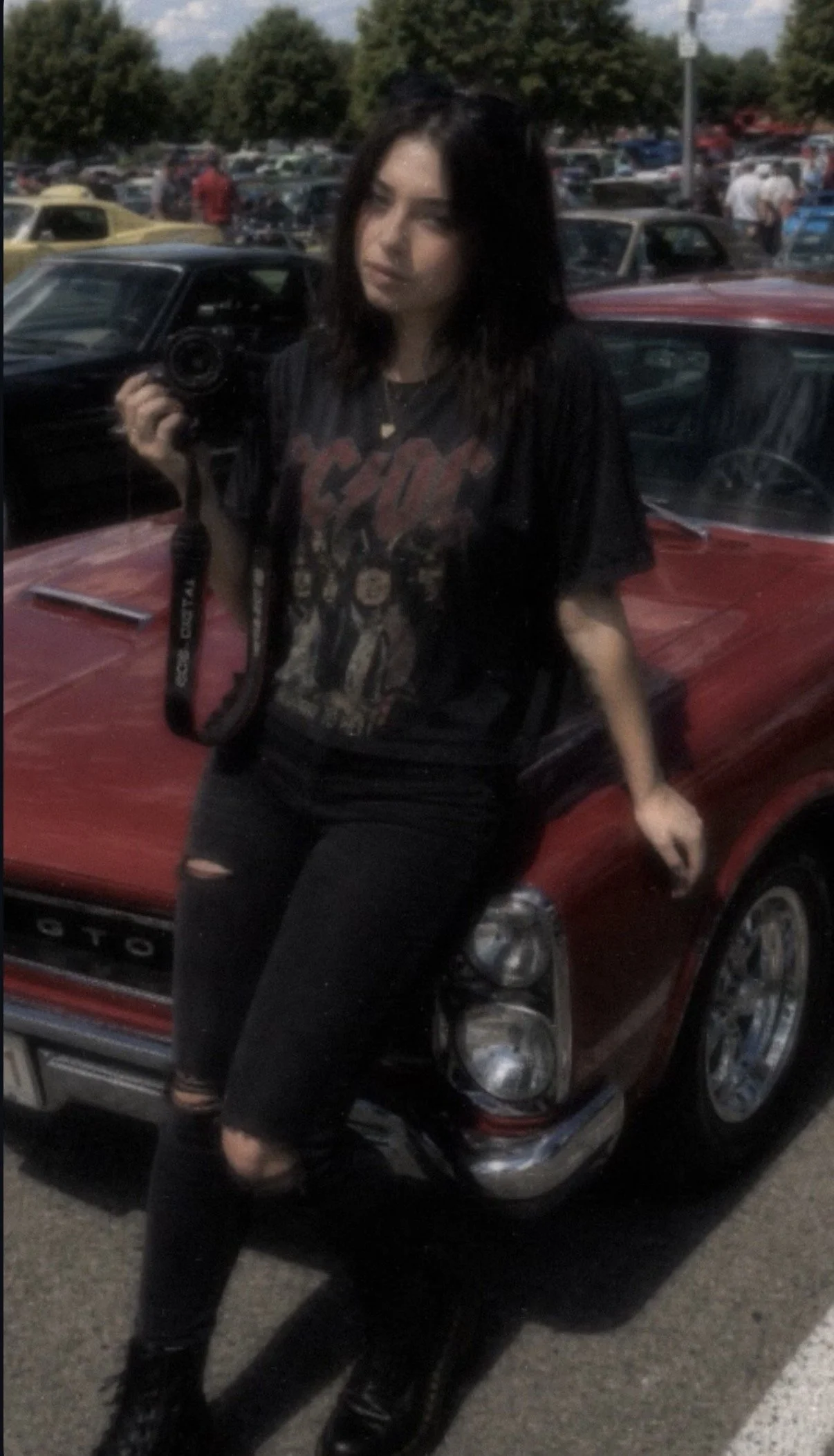 A young woman with dark, wavy hair standing in front of a red vintage car in a parking lot filled with old cars and trees in the background. She is holding a camera and wearing a black graphic t-shirt, ripped black jeans, and black boots.