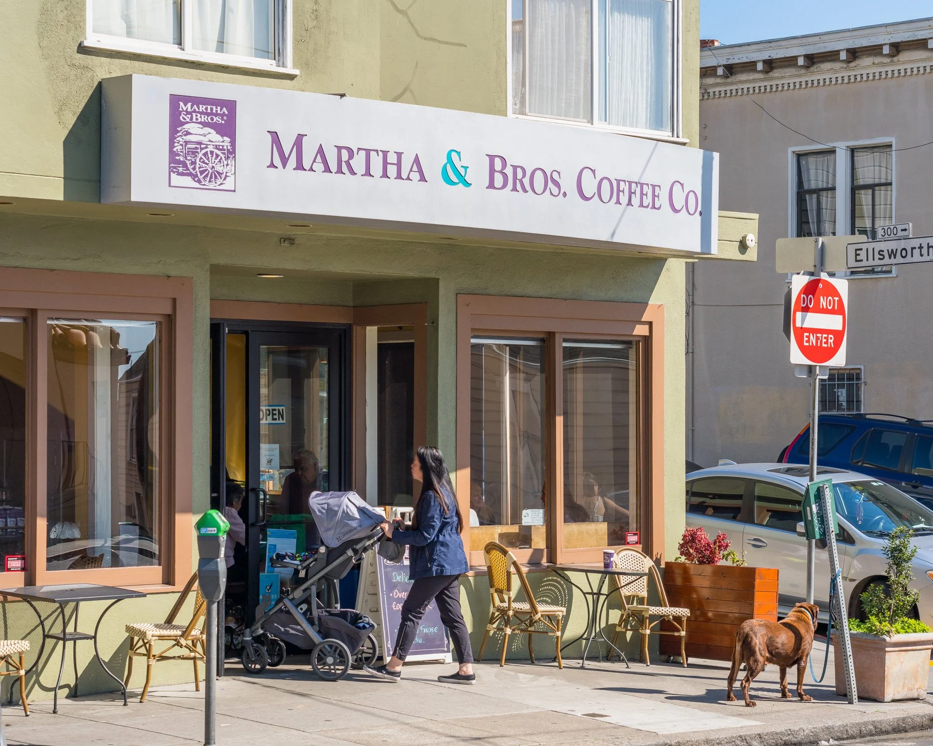 Exterior view of Martha & Bros. Coffee Co. storefront with outdoor seating, customers, and a dog. Sign with purple and blue text, and a logo with a wagon. People are entering the shop, and there are chairs and flowers outside.