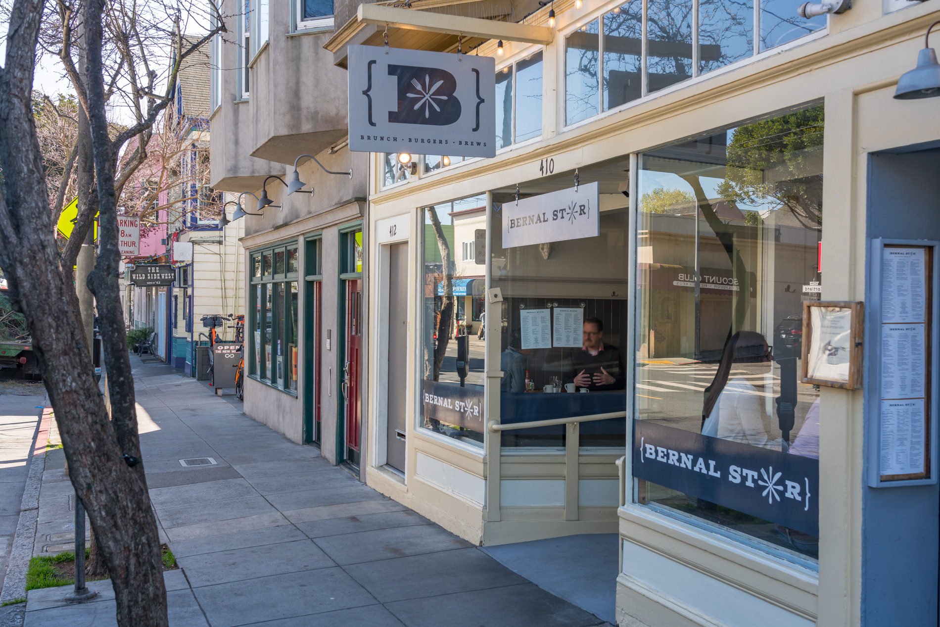 Street view of a restaurant on Bernal Street with large glass windows and white exterior, a man sitting inside near the window, and signs overhead indicating brunch, burgers, and brews.