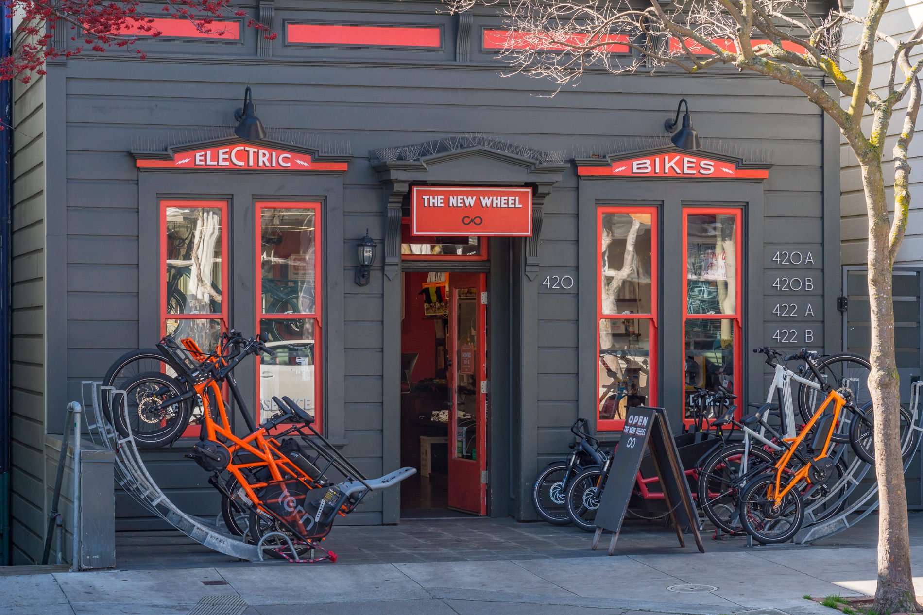 Front facade of a bicycle shop called 'The New Wheel' with a sign that reads 'Electric Bikes'. Store has large windows displaying bikes inside. Multiple bikes, including an orange electric bike, are parked outside and on a bike rack. A street tree wi