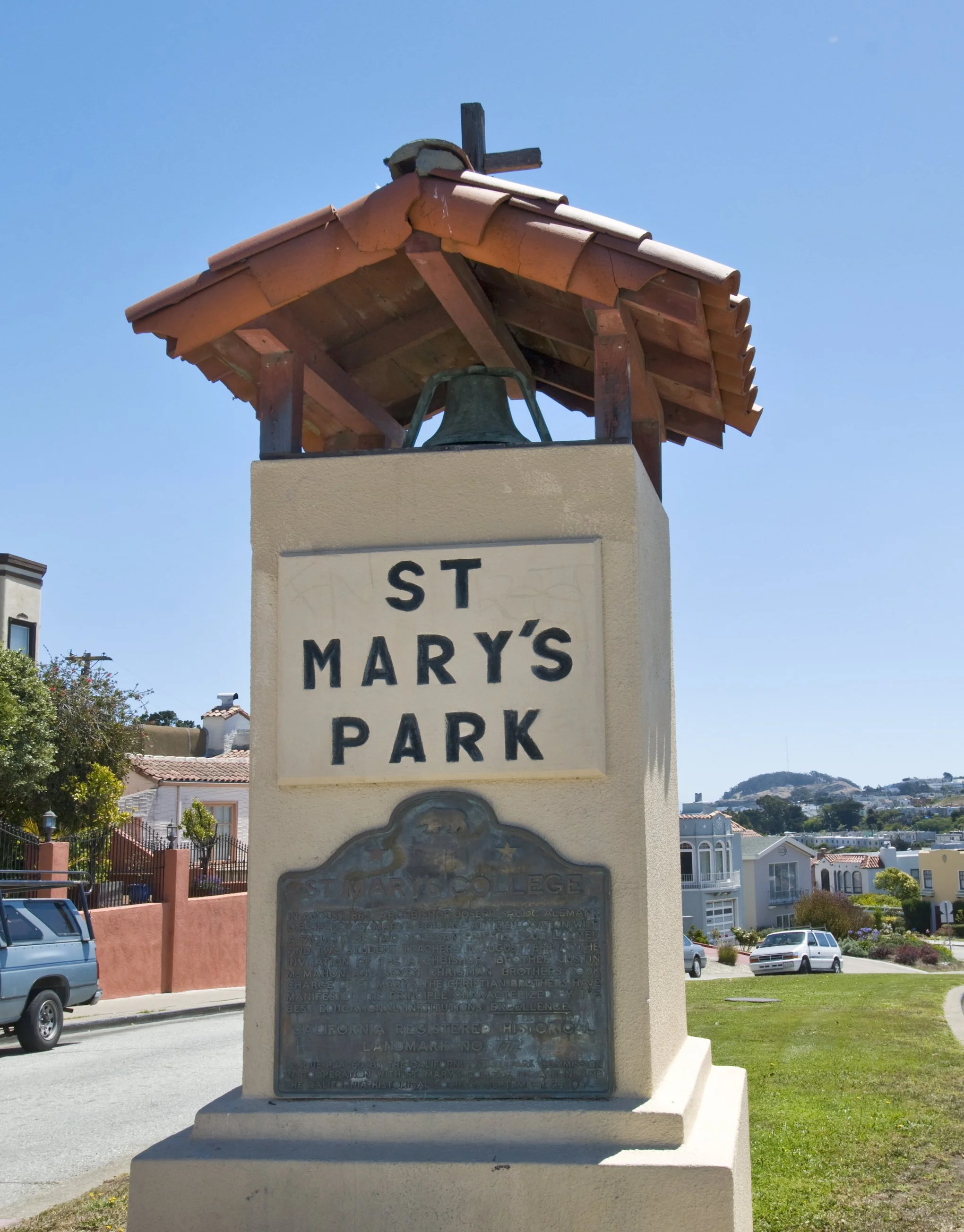A historic monument with a bell on a beige stone base, located in a park named St. Mary's Park in California, with a background of residential buildings and a hillside