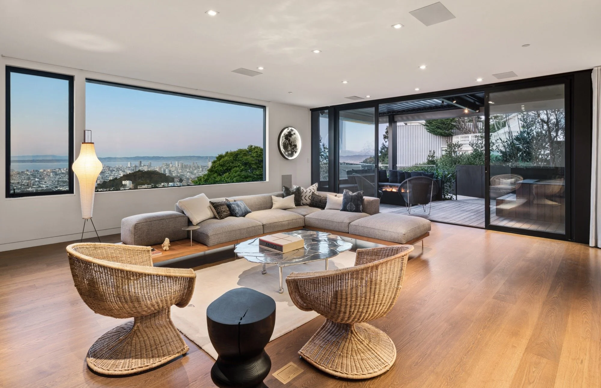 Living room with large window view of cityscape, beige sectional sofa with cushions, wicker chairs, marble coffee table, floor lamp, sliding glass doors leading to outdoor patio with furniture and plants.