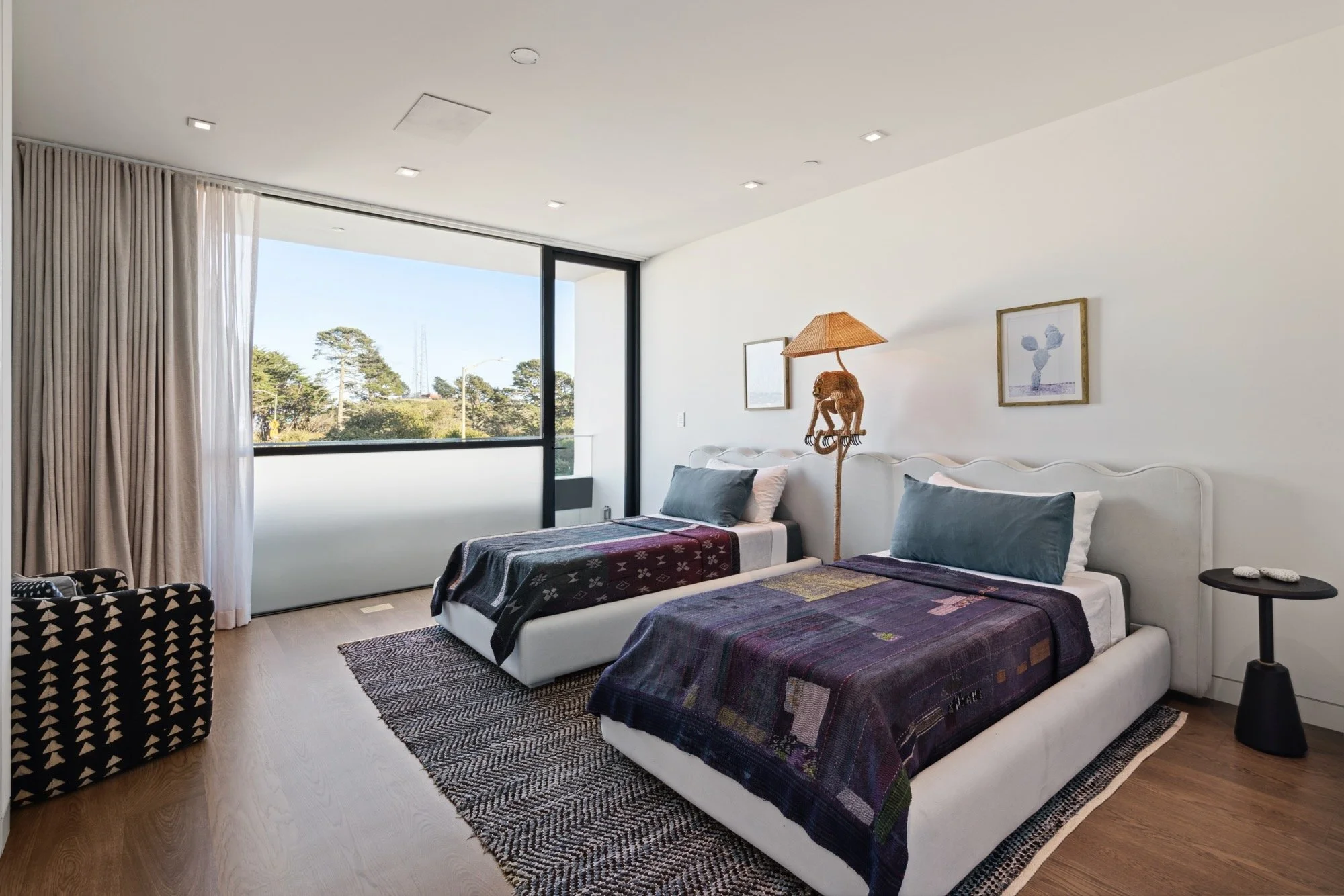 A modern bedroom with two beds, each with colorful blankets and gray pillows, a black side table with white stones, a decorative plant lamp, framed artwork, a large window with curtains, and a partial view of outside trees.