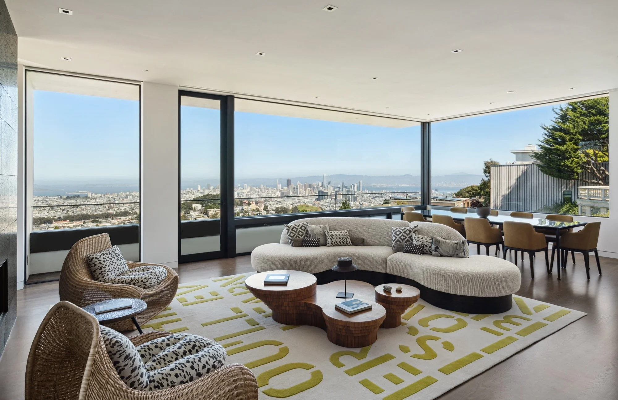 Modern living room with large windows offering a city skyline view, contemporary furniture including a curved sofa, patterned armchairs with pillows, and a wooden coffee table on a yellow and white geometric rug.