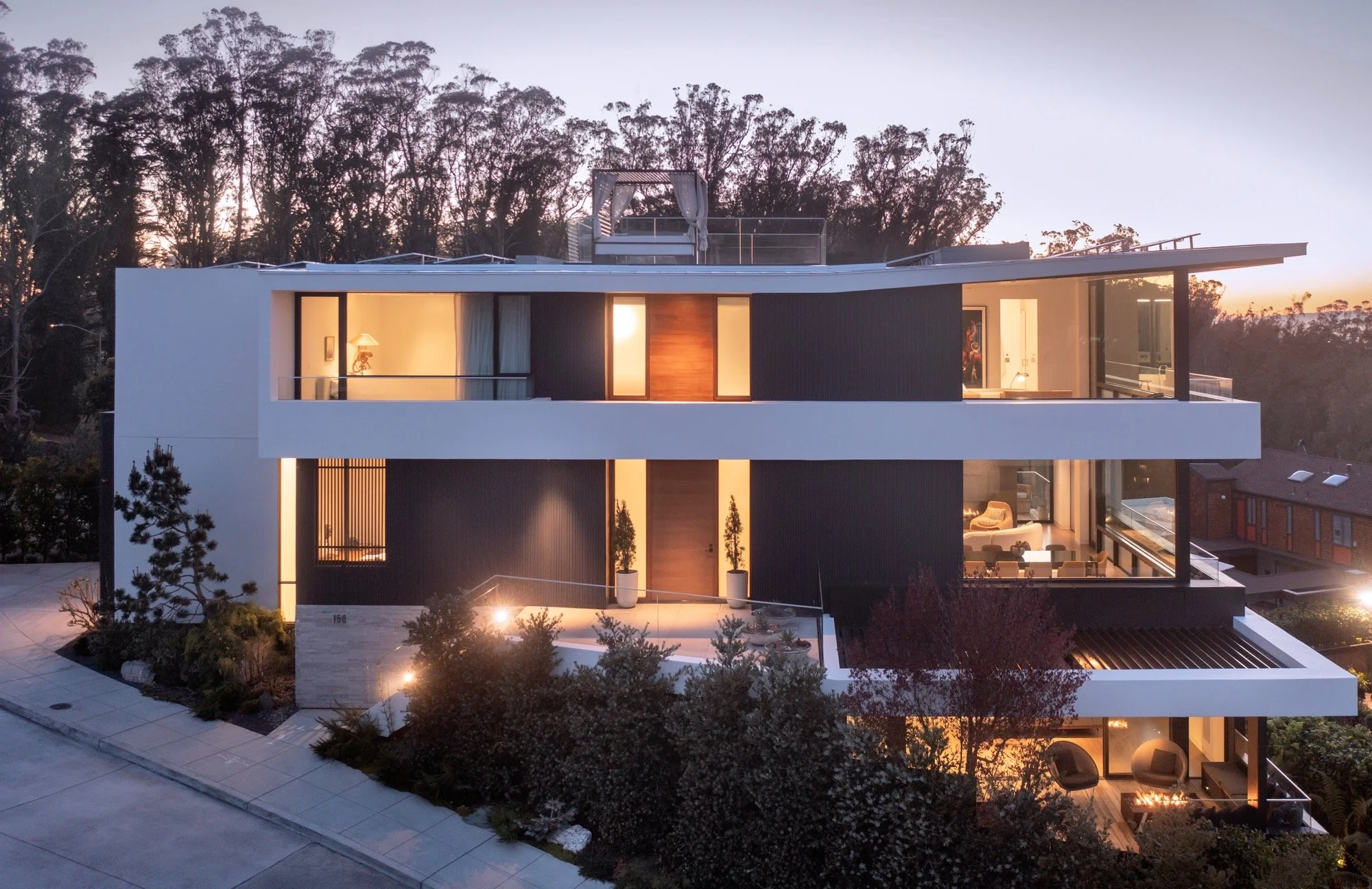Modern multi-story house at dusk with illuminated interior, large glass windows, and a rooftop terrace surrounded by trees.
