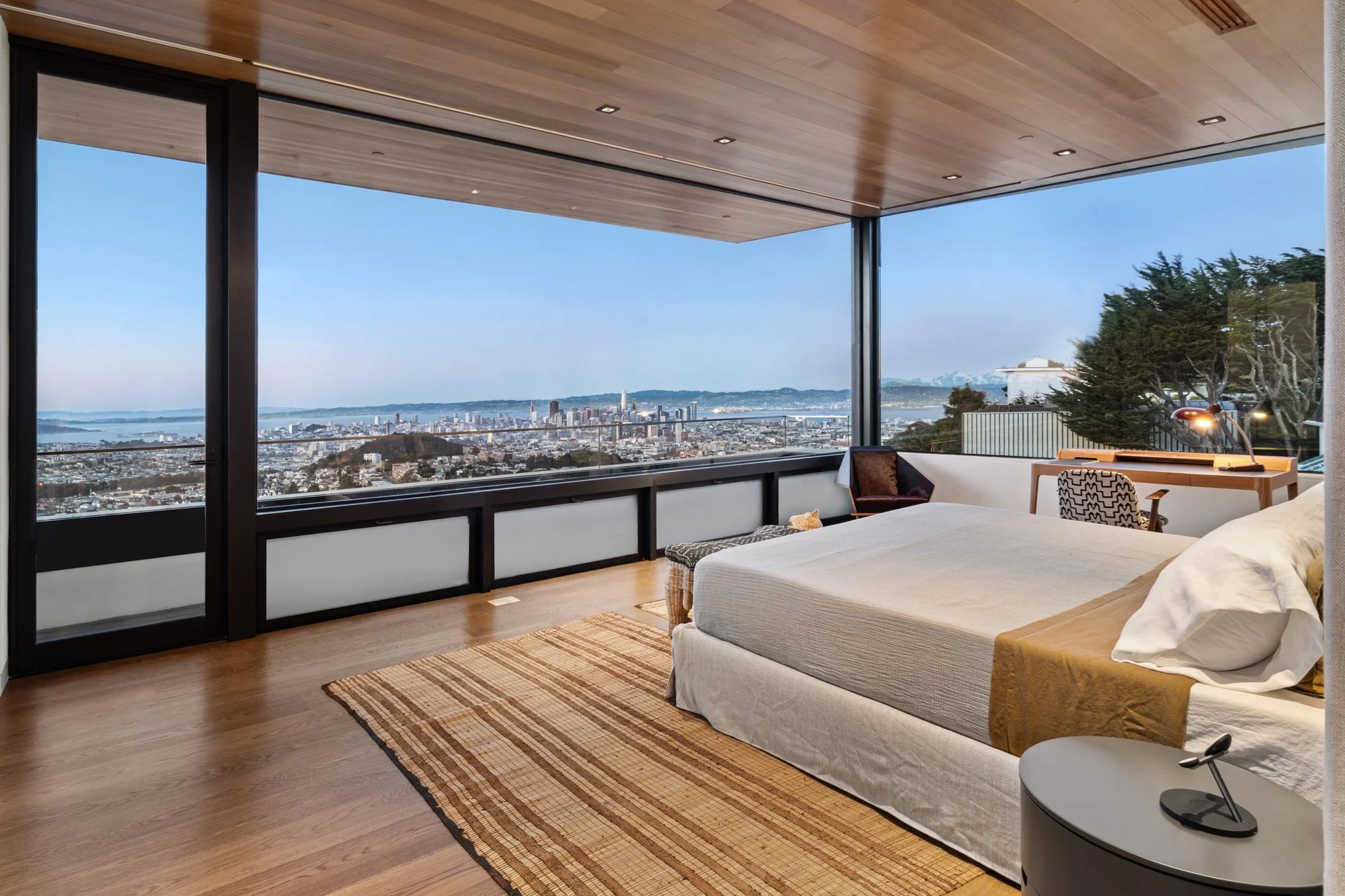 A modern bedroom with a large window offering a view of a city and a bay, a bed with a white cover, a patterned rug, a small side table, a desk with a chair, and a wood-paneled ceiling with recessed lighting.