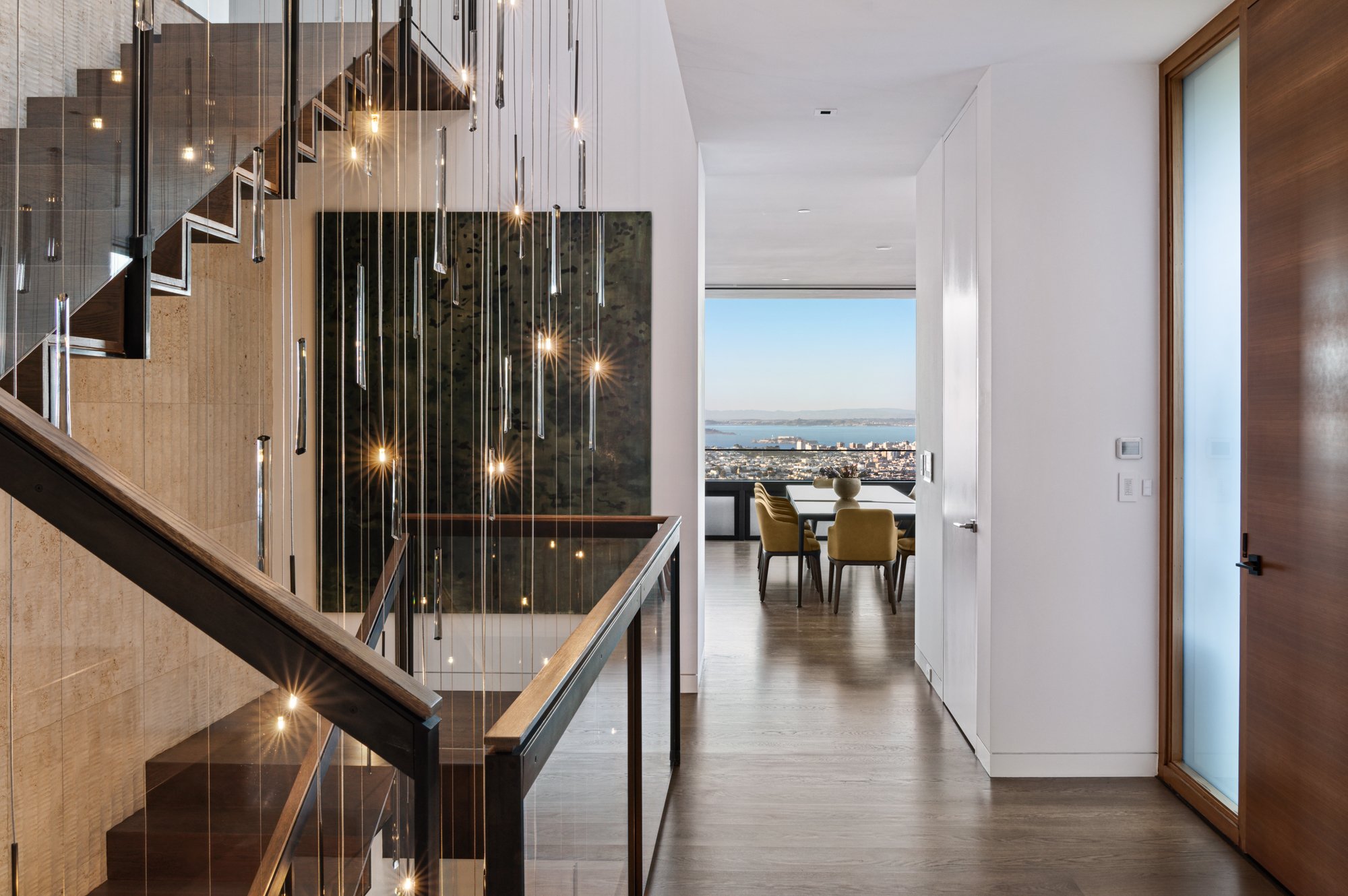 Modern interior with staircase featuring glass railings and hanging pendant lights, a dining area with a city view through large windows, and a door on the right side.