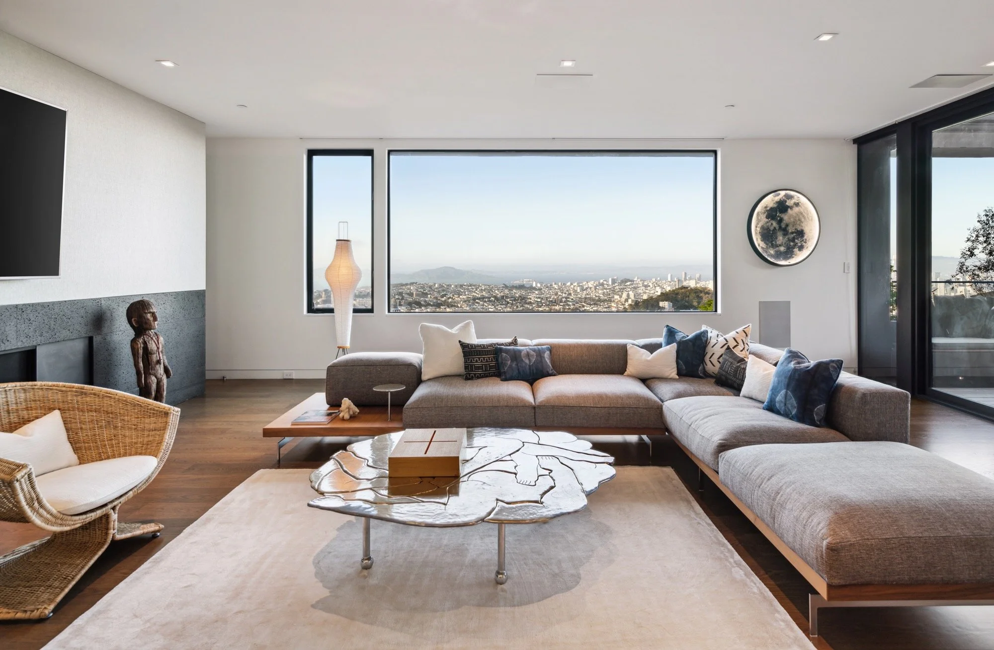 Living room with large windows showing city and mountain view, beige sectional sofa with pillows, white marble coffee table, wicker chair with cushion, and a moon decorative wall hanging.