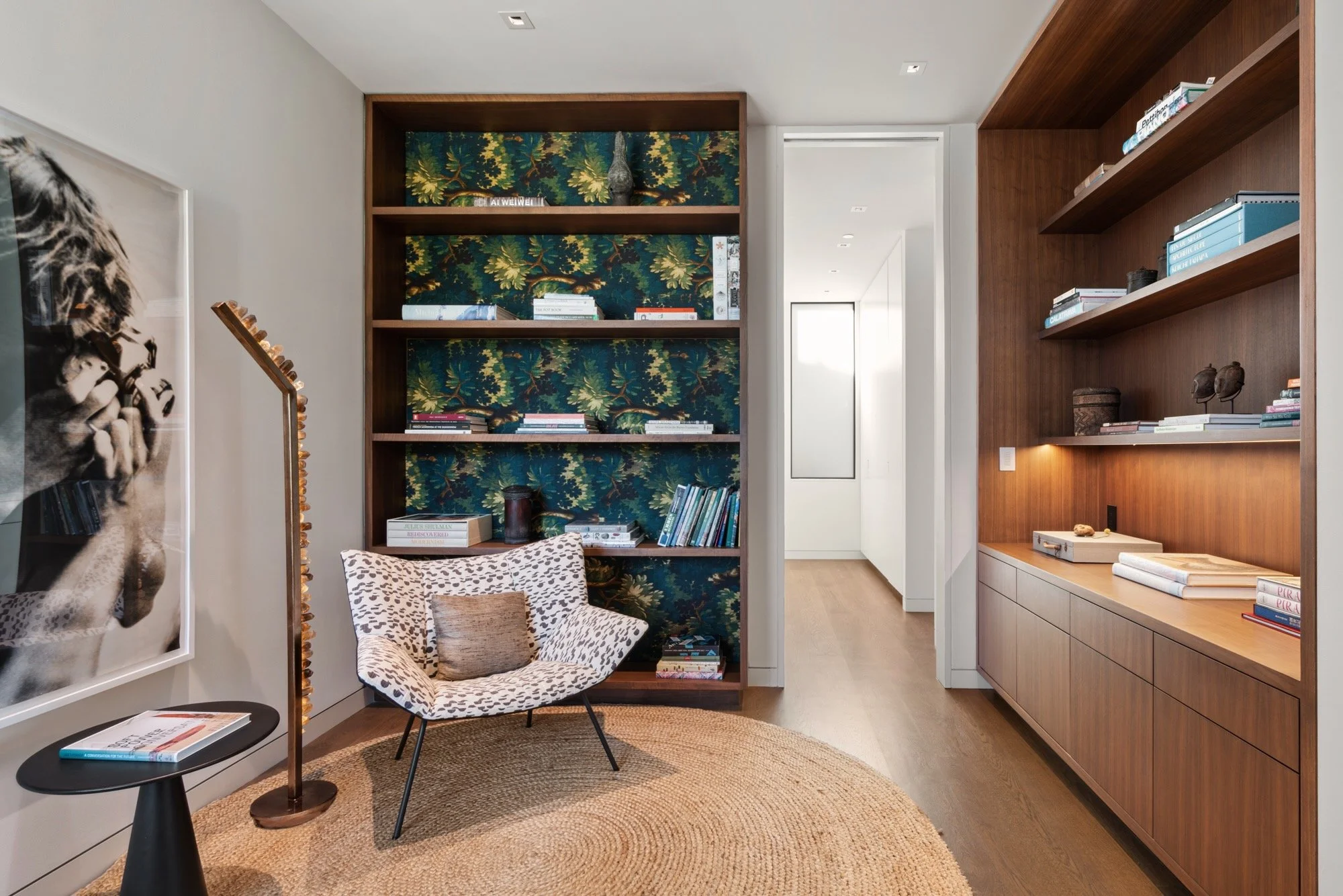 Interior of a modern living space featuring a round woven rug, a patterned accent chair, built-in bookshelves with books and decorative items, and framing a hallway with a doorway and ceiling lights.