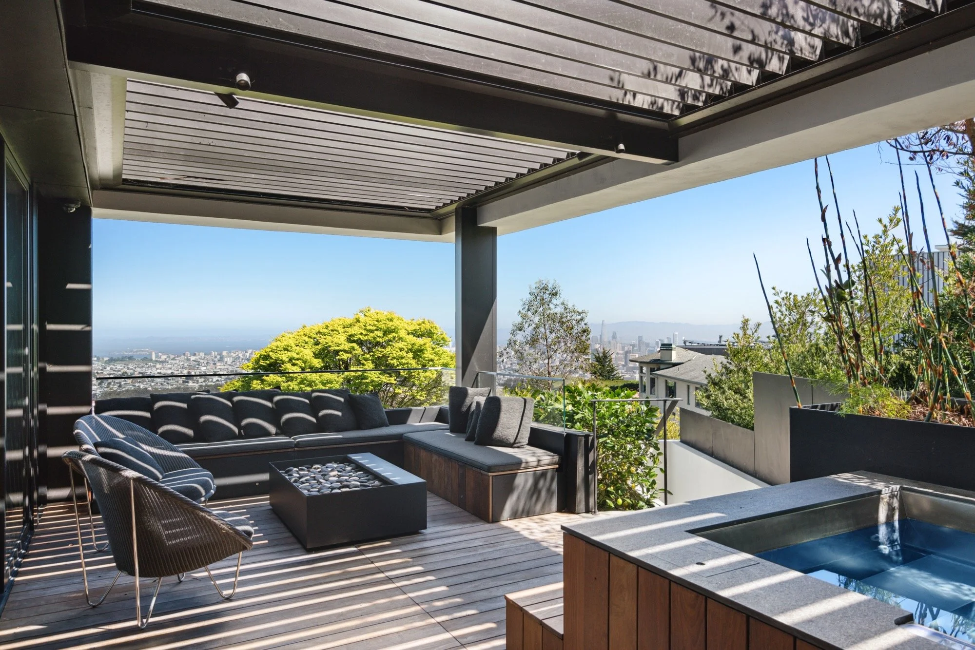 Outdoor patio with black modern furniture, a hot tub, and lush green trees in a high-rise setting with city skyline view