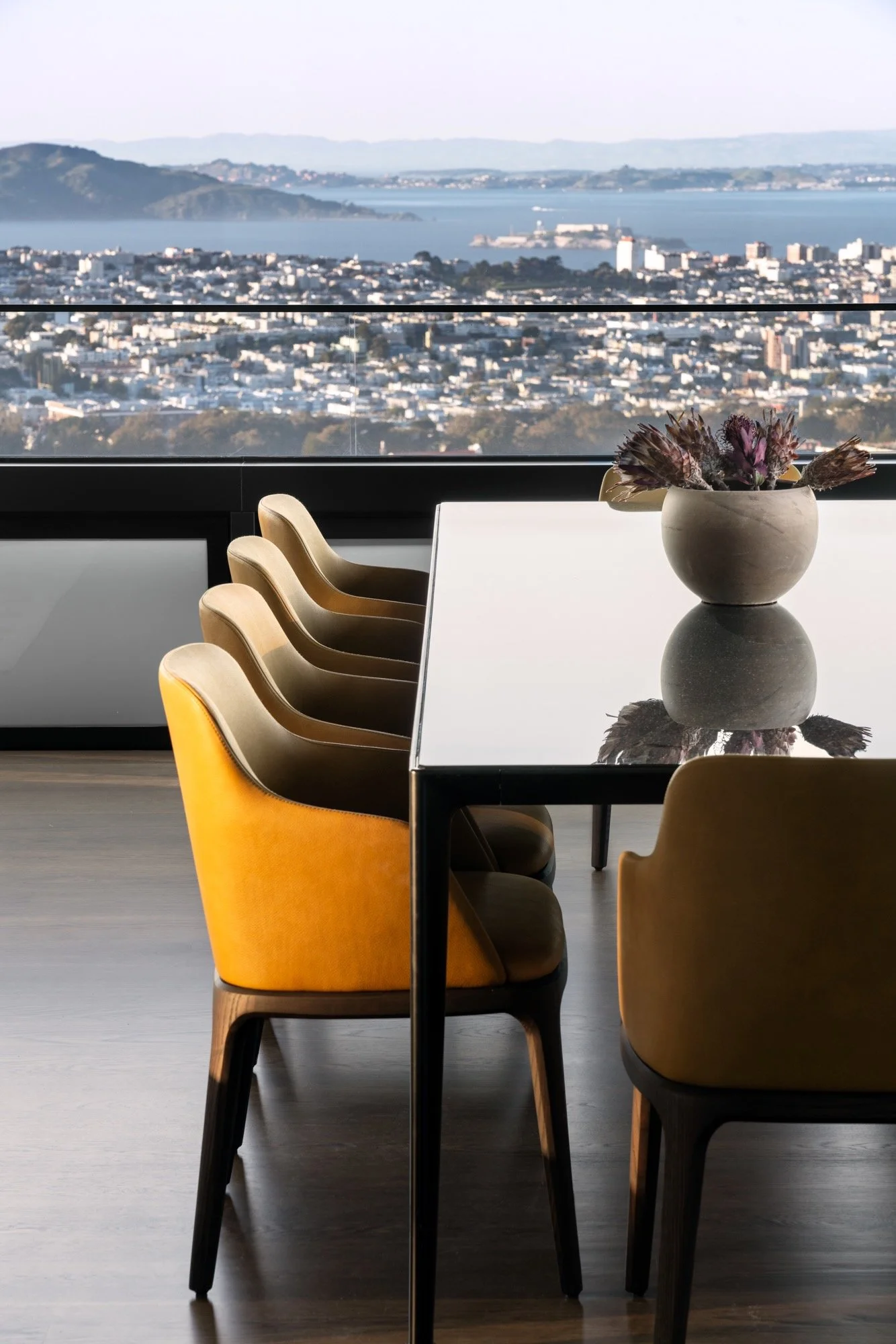 Modern dining room with a white table and yellow upholstered chairs, large windows revealing a cityscape and water view with distant hills.
