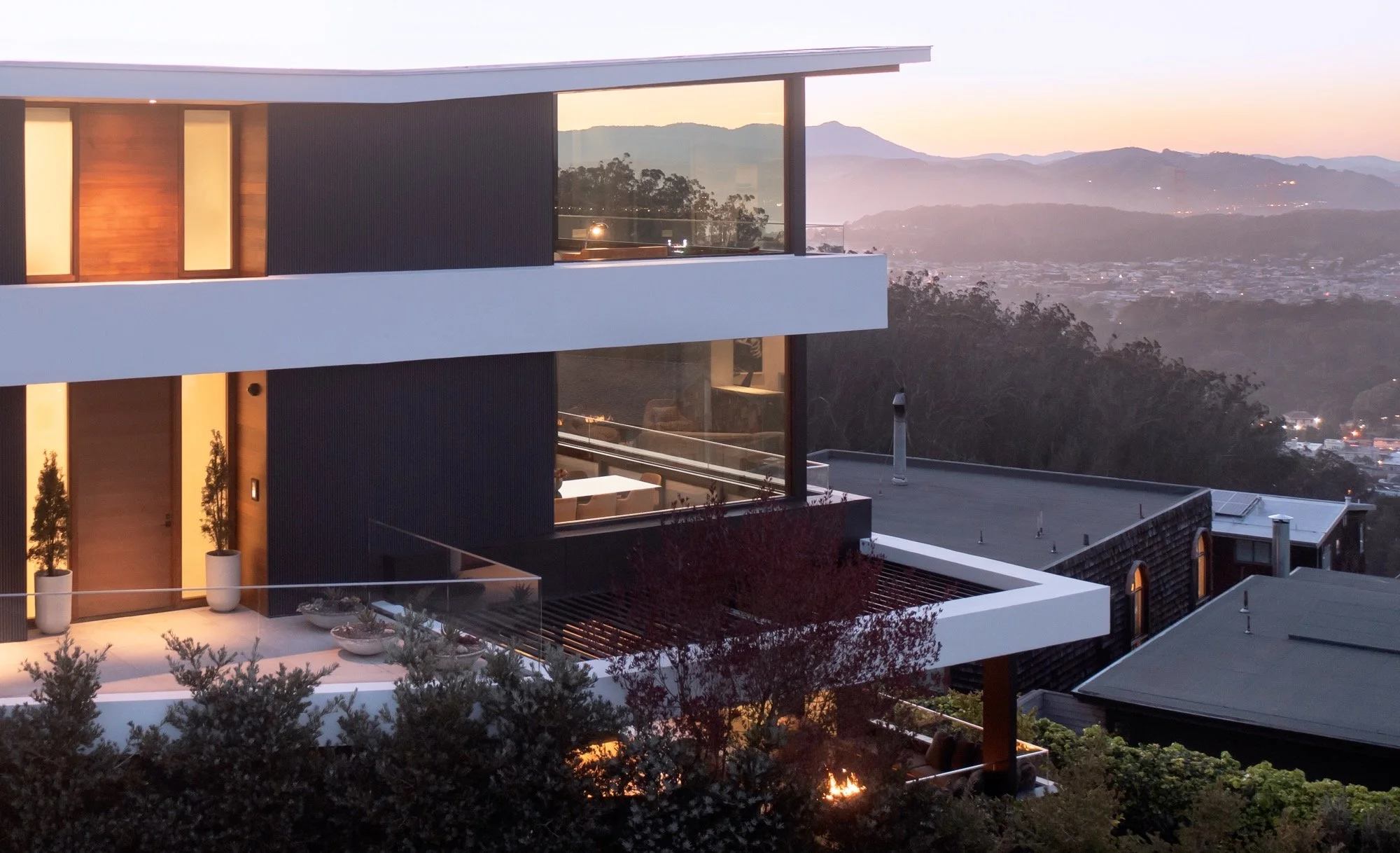 Modern multi-story house with large glass windows overlooking a mountain range at sunset, surrounded by trees and shrubs.