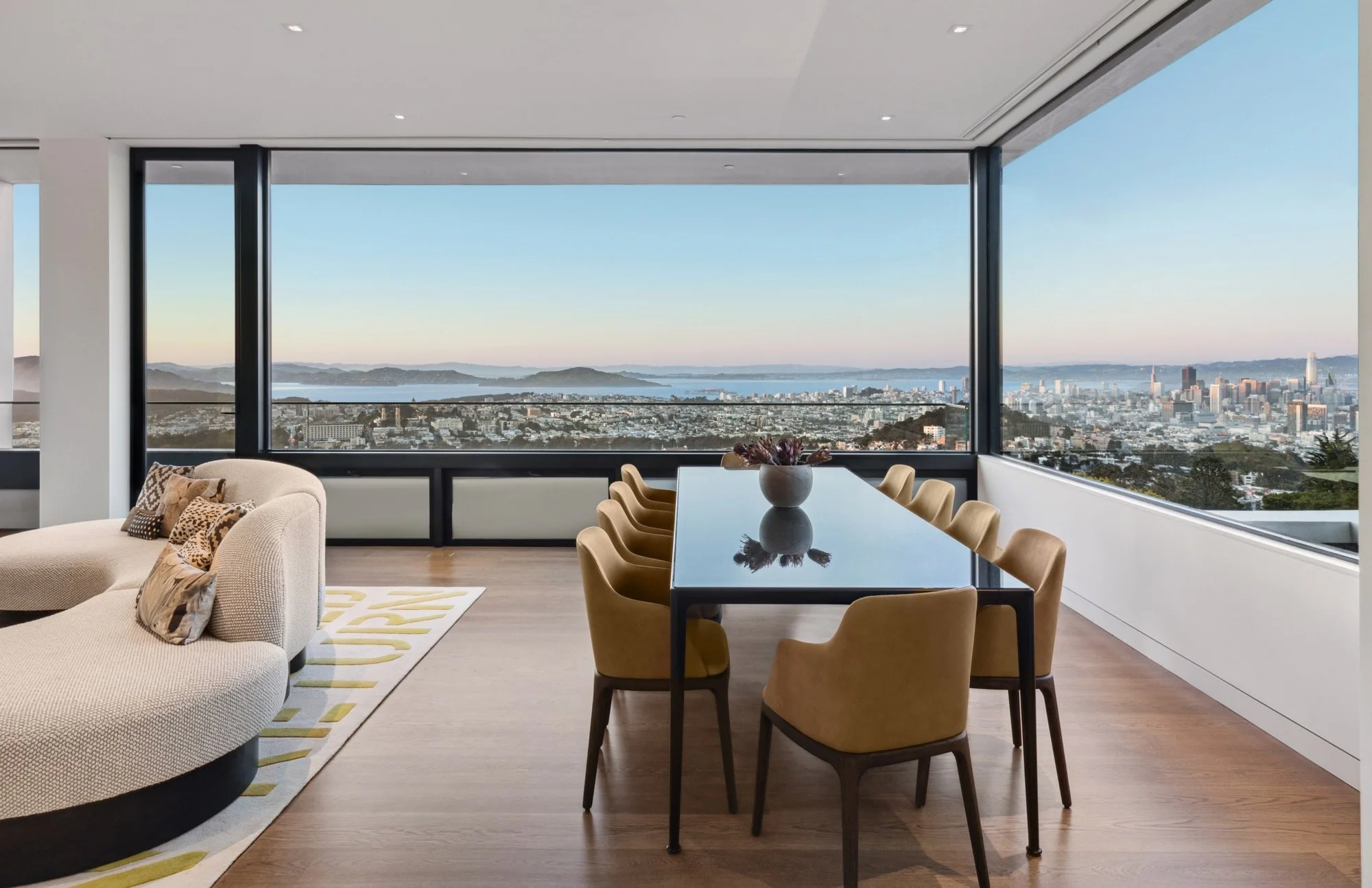 Modern dining and living room with large windows overlooking city and water view, beige sofa with patterned pillows, wooden dining table with yellow chairs, decorative vase with flowers.