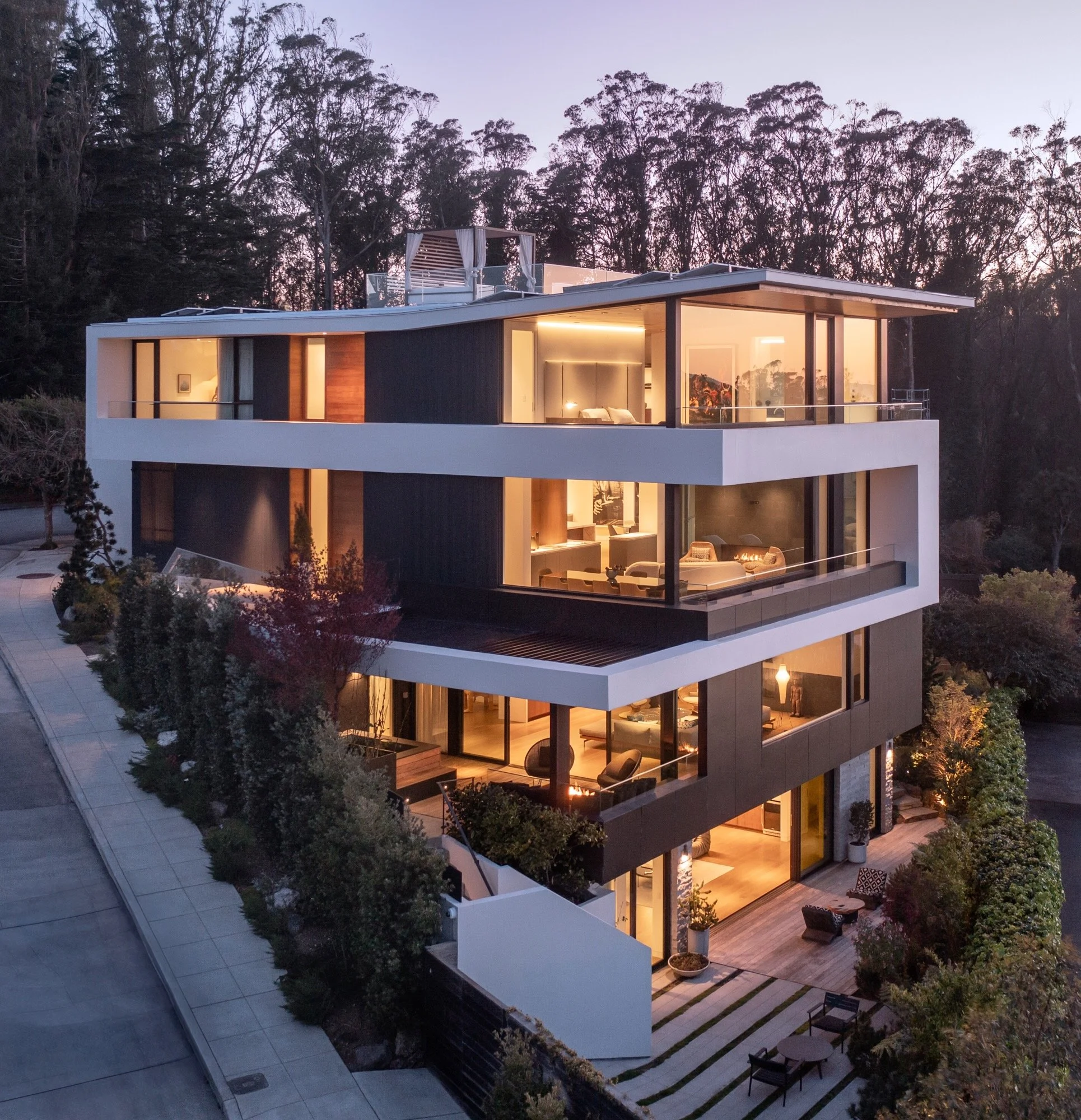 Modern multi-story house with large glass windows illuminated from inside, set against a background of trees at dusk. The house has outdoor seating and landscaped steps. Interior rooms and outdoor patio furniture are visible.