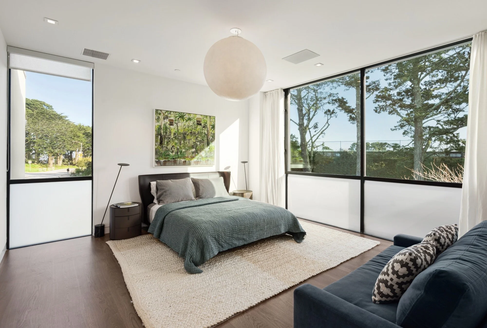 Modern bedroom with large windows, a bed with gray bedding, a textured beige rug, a navy blue armchair with patterned pillows, and artwork on the wall, illuminated by natural light.
