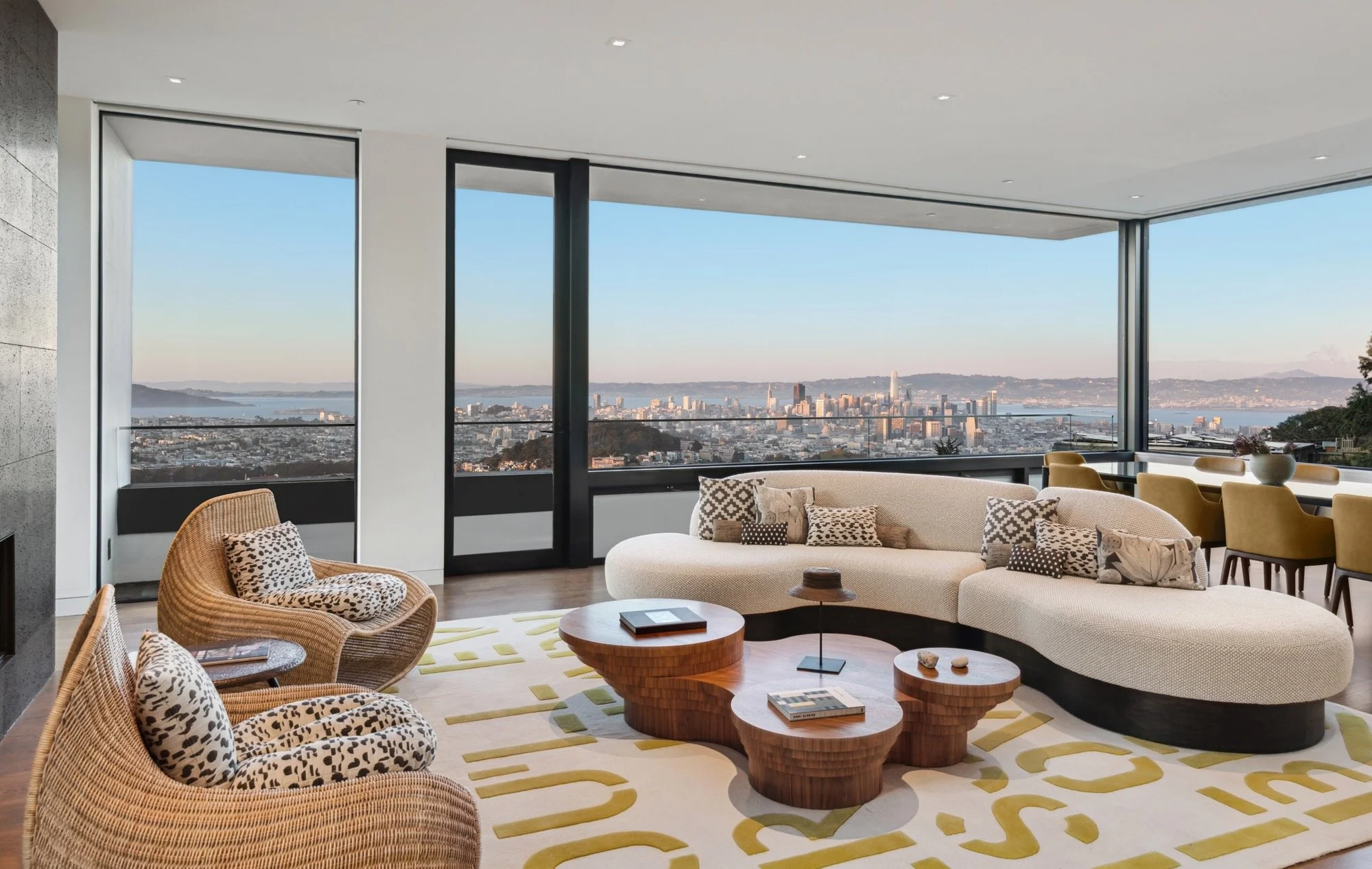 Modern living room with large windows overlooking a cityscape and water. The room features a curved beige sofa with assorted patterned pillows, a round wooden coffee table, and wicker chairs with leopard print cushions. A yellow dining chair is visib