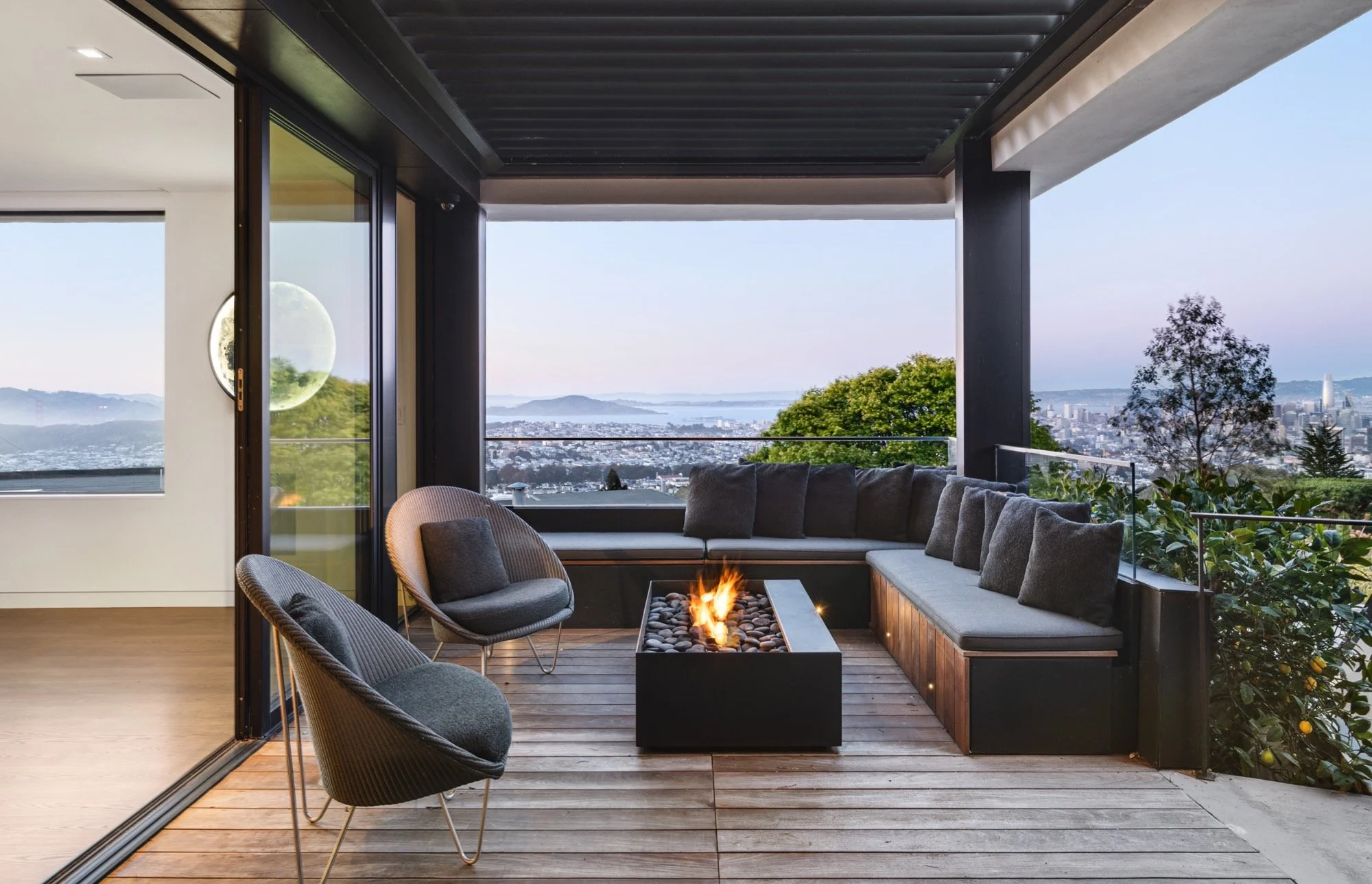 Outdoor balcony with a firepit table, cushioned seating, and glass railing overlooking a cityscape with hills and water in the background.