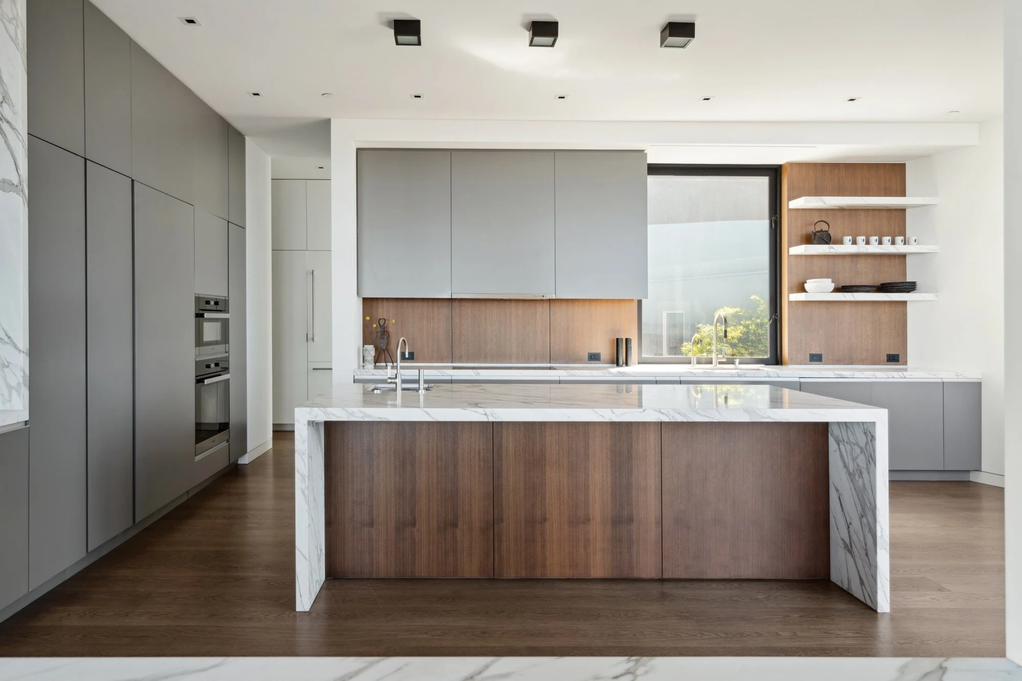 Modern kitchen with wood and marble accents, gray cabinets, open shelving, and a large window with a view of trees.
