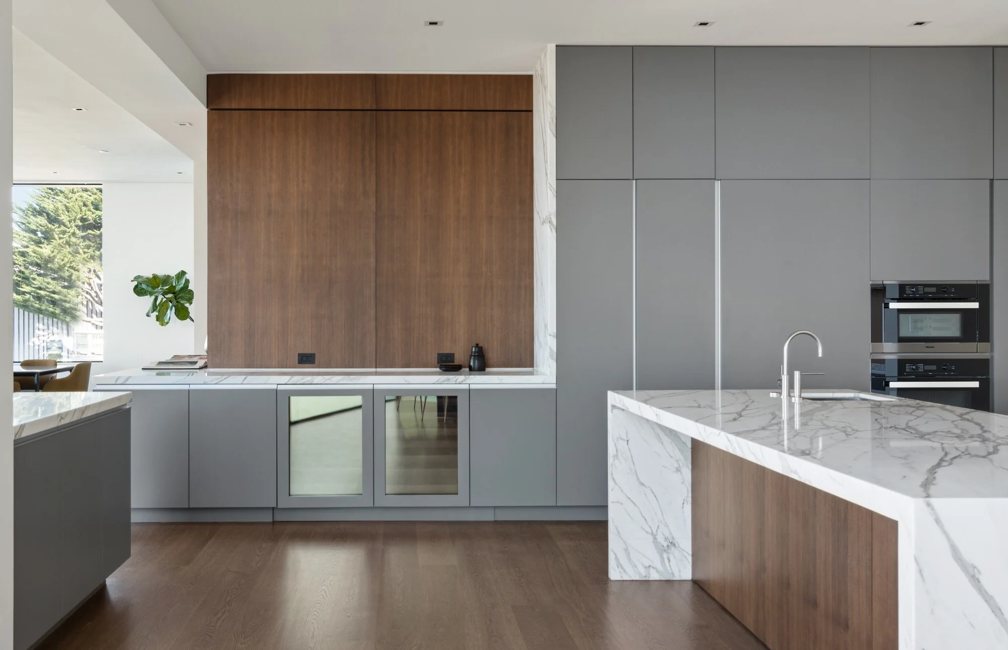 Modern kitchen with white marble countertops, gray cabinets, a wooden accent wall, built-in ovens, and a large window with a view of trees.