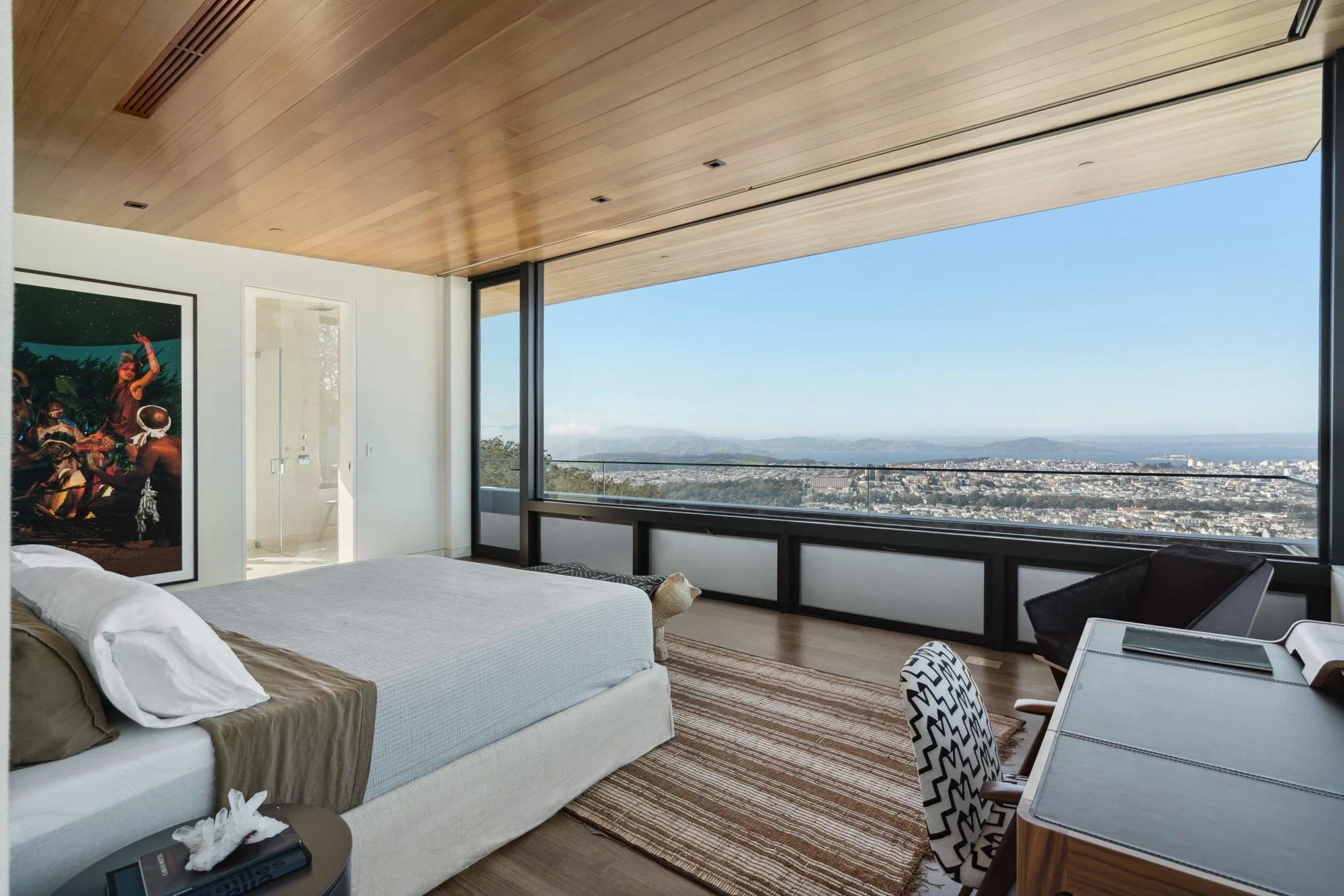A bedroom with a large window showing a cityscape and mountains in the distance, a bed with white and beige bedding, a framed artwork, and a desk with a patterned chair.