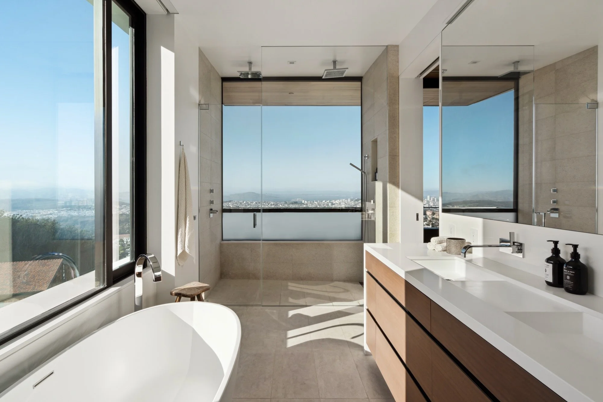 Modern bathroom with large windows showing city views, a white bathtub, a wooden vanity with black fixtures, and a walk-in shower with glass doors.