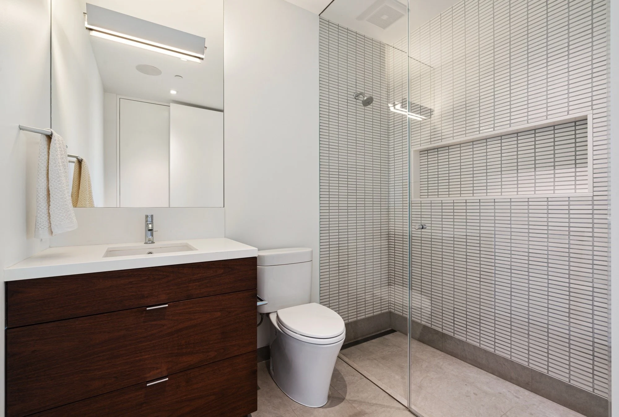 Modern bathroom featuring a wooden vanity with white sink, a mirror, a toilet, and a glass-enclosed shower with white subway tiles.