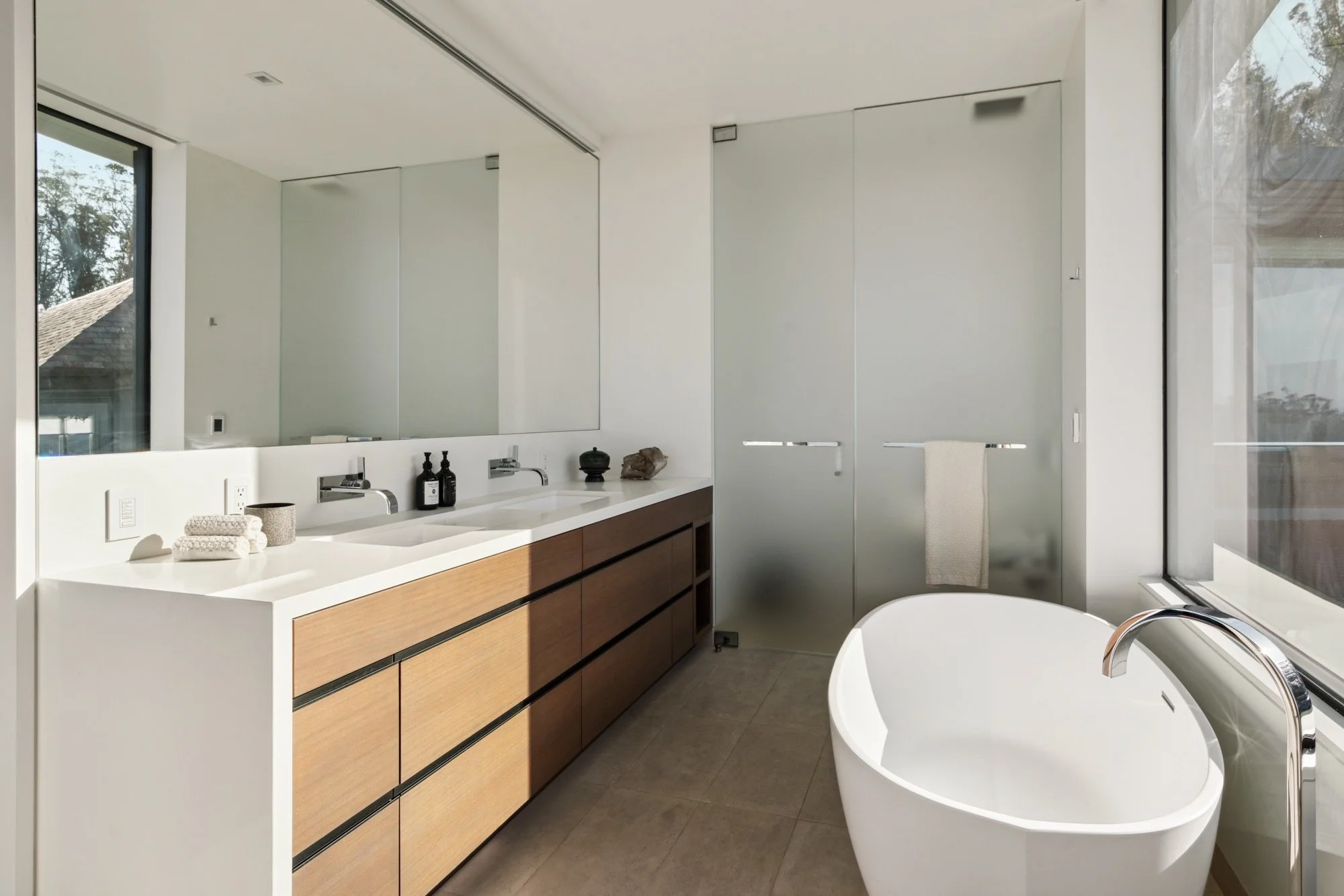 Modern bathroom with a white vanity, wooden cabinet drawers, and a freestanding oval bathtub near large windows.