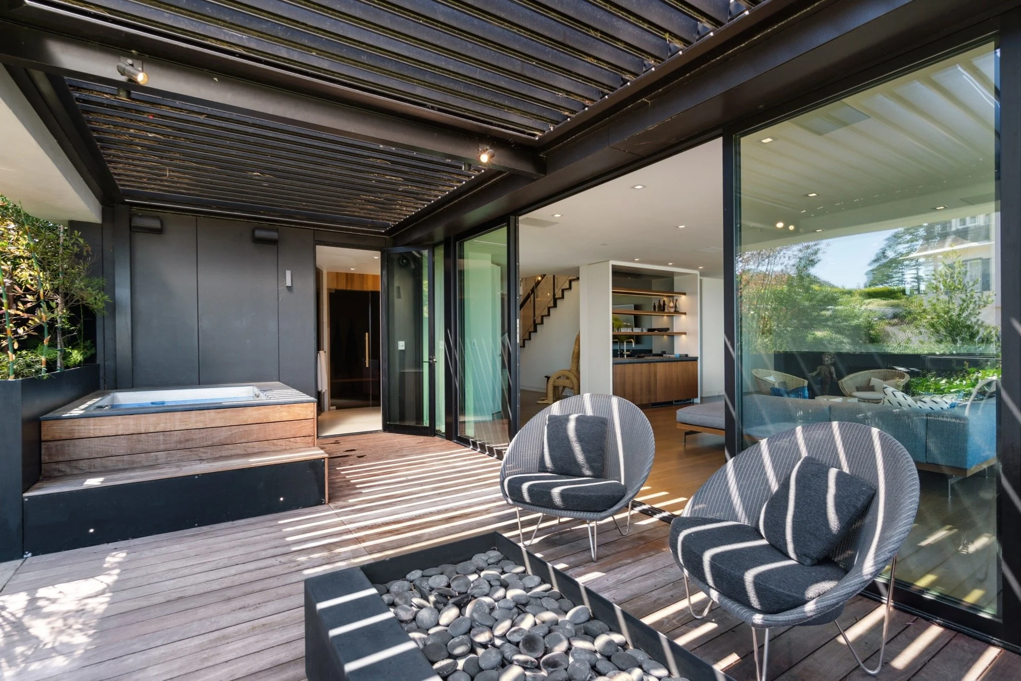 A modern outdoor patio area with a hot tub, a round chair, and a fire pit filled with black stones. The patio has a wooden deck and is partially covered by a slatted pergola. The interior of the house is visible through large glass sliding doors, sho