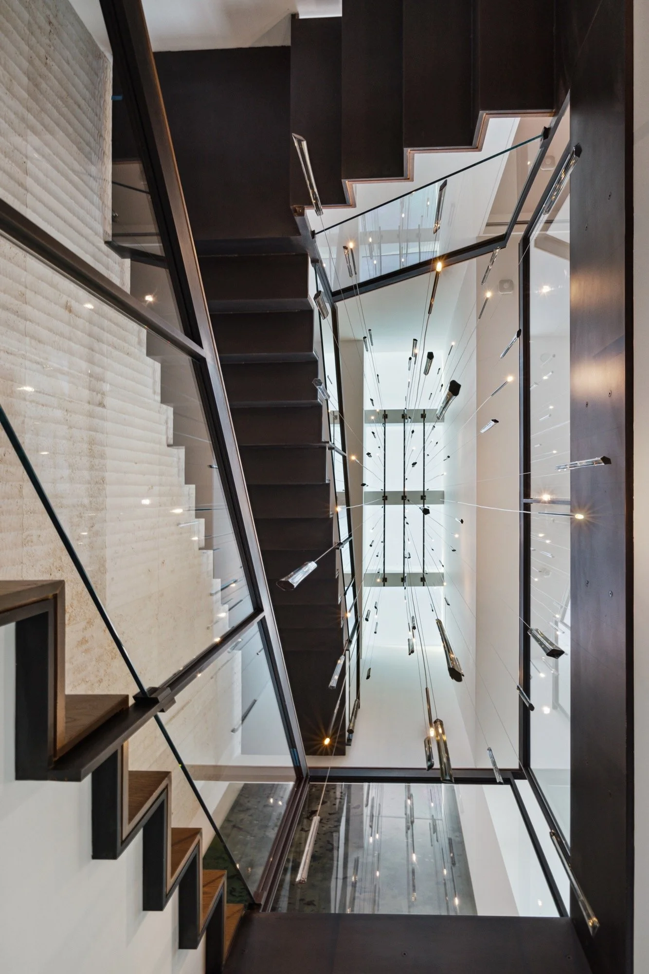 A modern staircase with glass railings and black steps, viewed from below, with a hanging light fixture and large windows in the background.