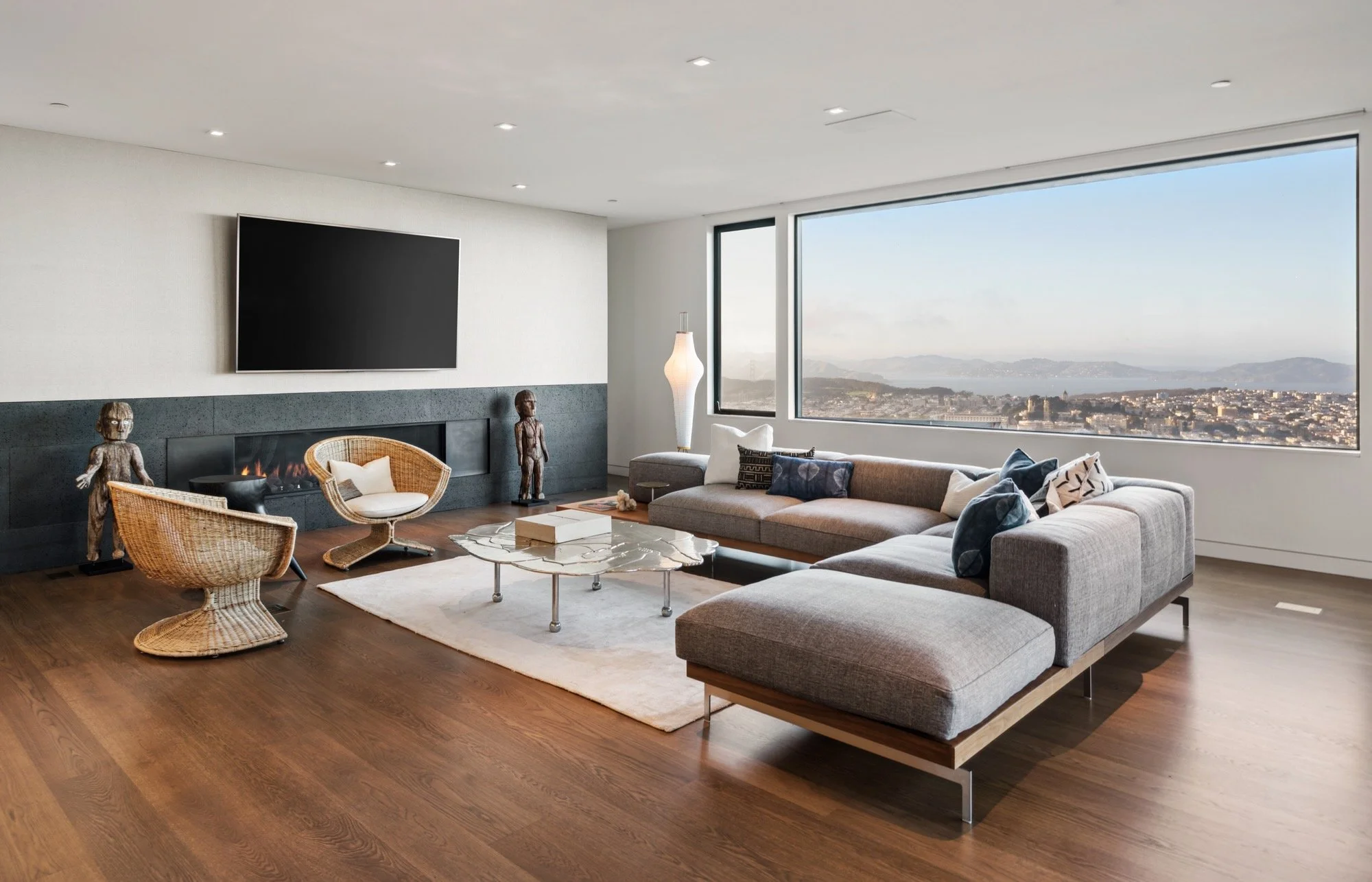 Modern living room with large windows overlooking cityscape, featuring a sectional sofa, rattan chairs, a marble coffee table, decorative statues, a fireplace, and a large flat-screen TV mounted on the wall.