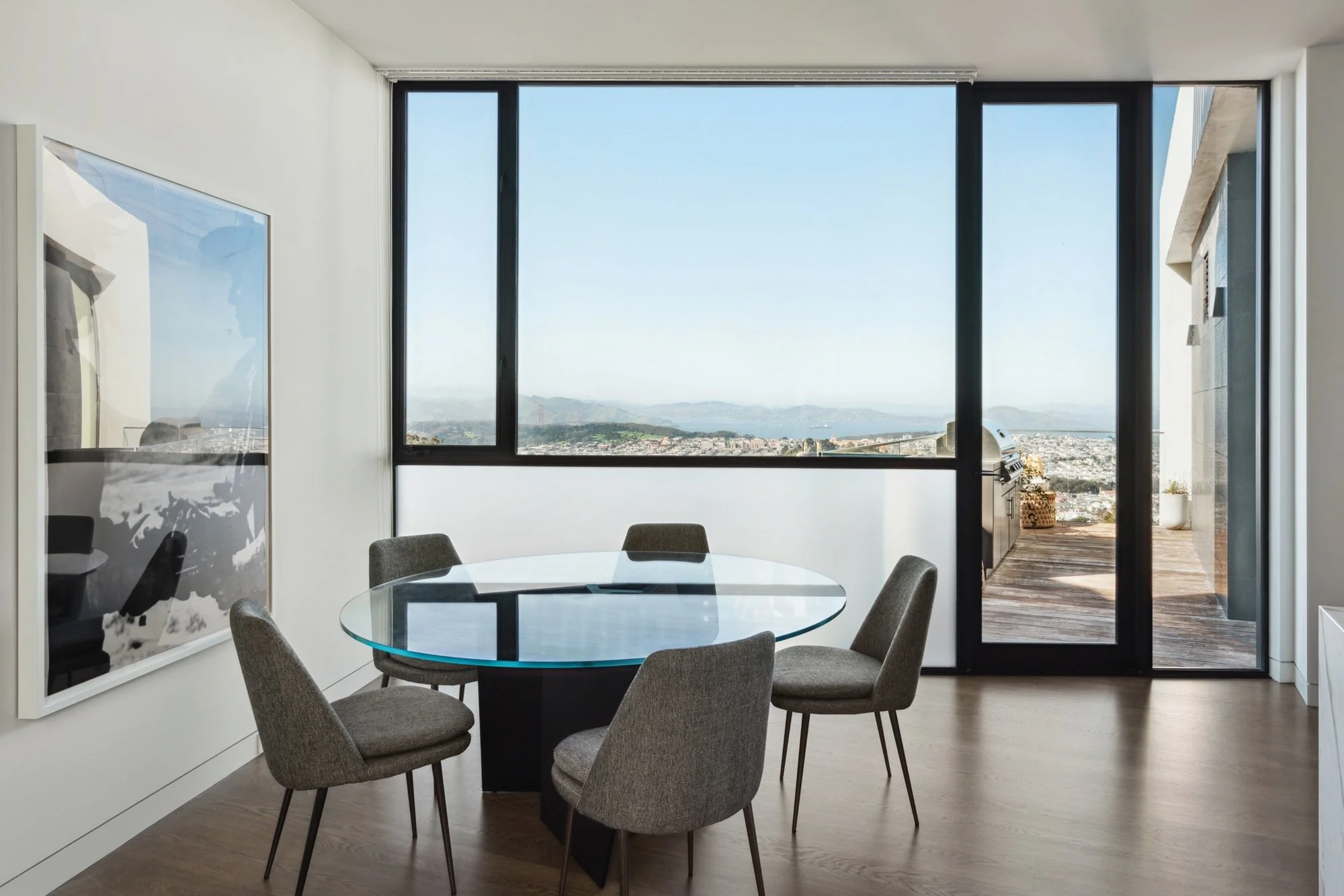 Modern dining area with a glass round table and six gray upholstered chairs. Large window with a view of distant hills and water, and a sliding door leading to an outdoor balcony with wooden flooring. Abstract art on the left wall.