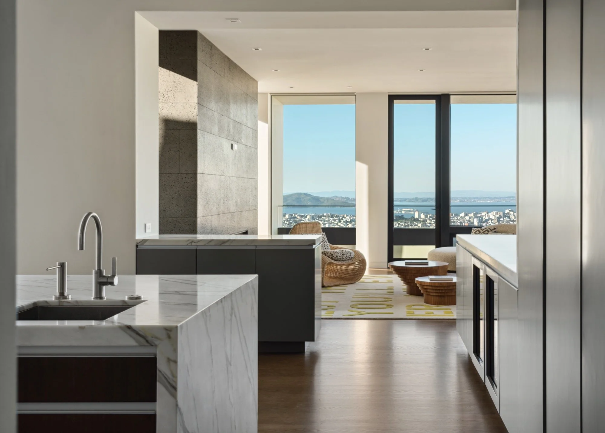 Modern kitchen with marble island, open living space with seating, large windows overlooking city skyline and mountains, minimalist decor, hardwood floors.