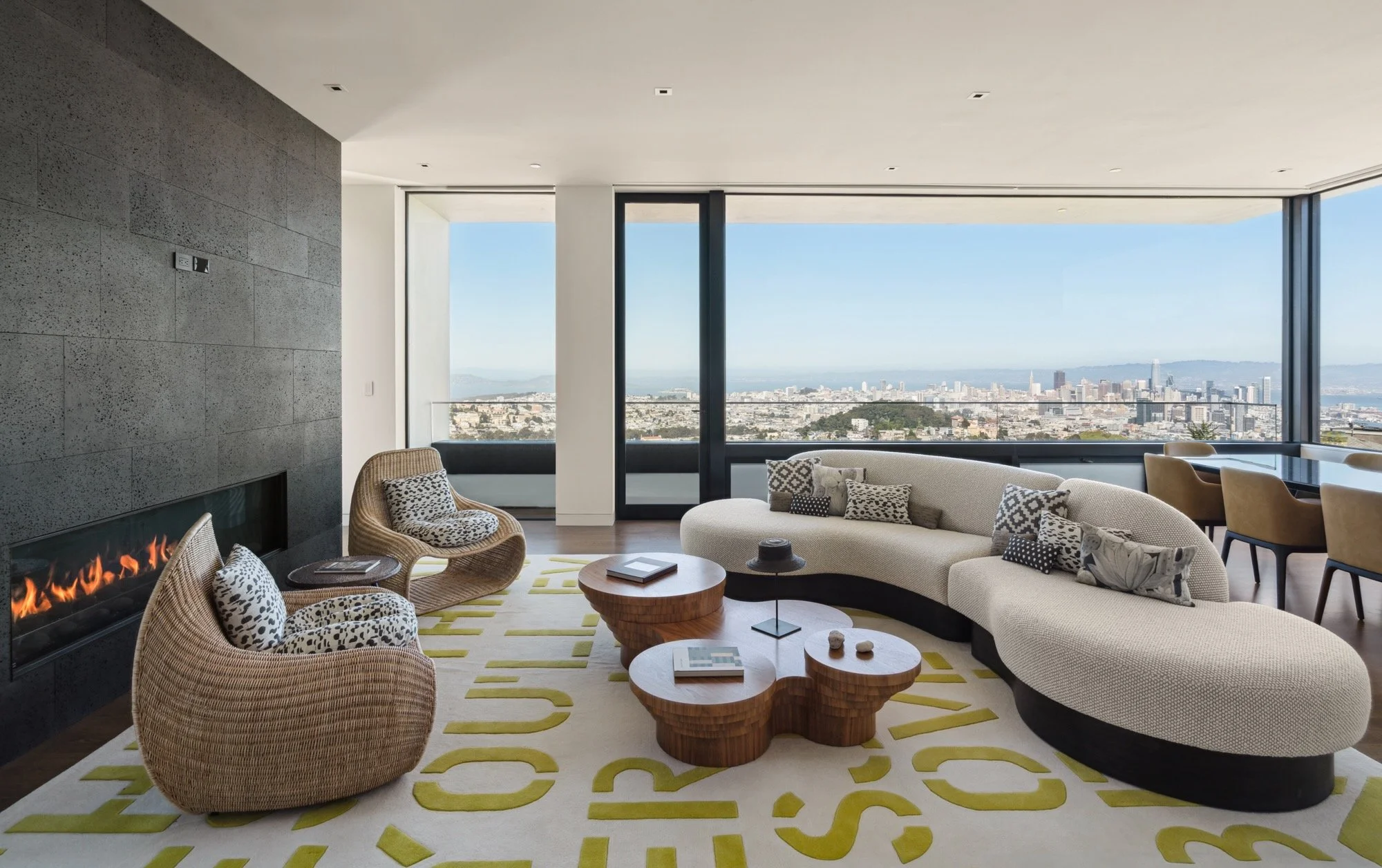 Modern living room with large windows showcasing a city view, curved sofa with patterned pillows, rattan armchair, fireplace, wooden coffee tables, yellow and white rug, and dining area with chairs.