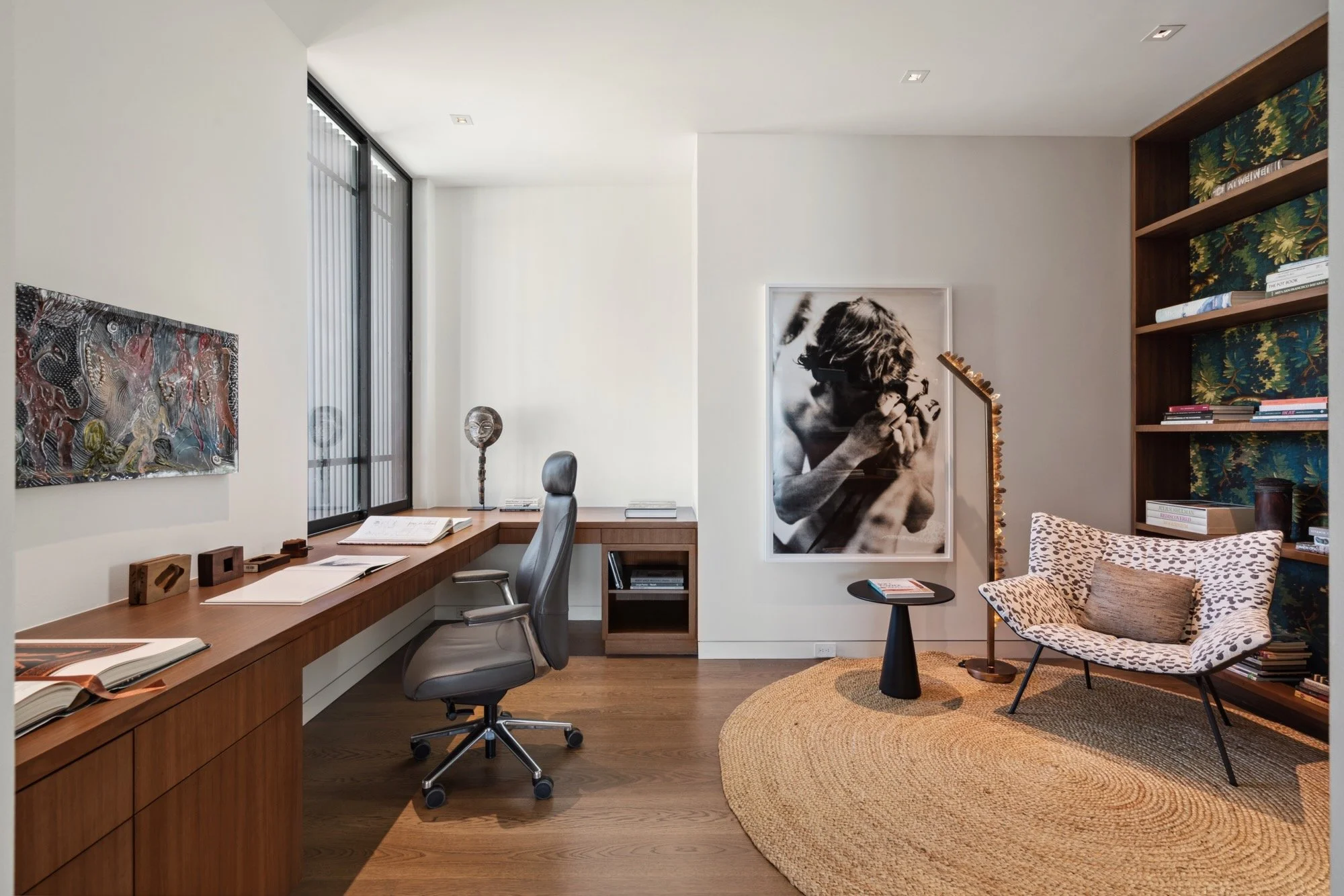 A modern home office with a large wooden desk, a gray office chair, and wall art including a black-and-white photo of a woman with curly hair playing a guitar. There's a wooden bookshelf filled with books, patterned wallpaper, and a cozy chair with c