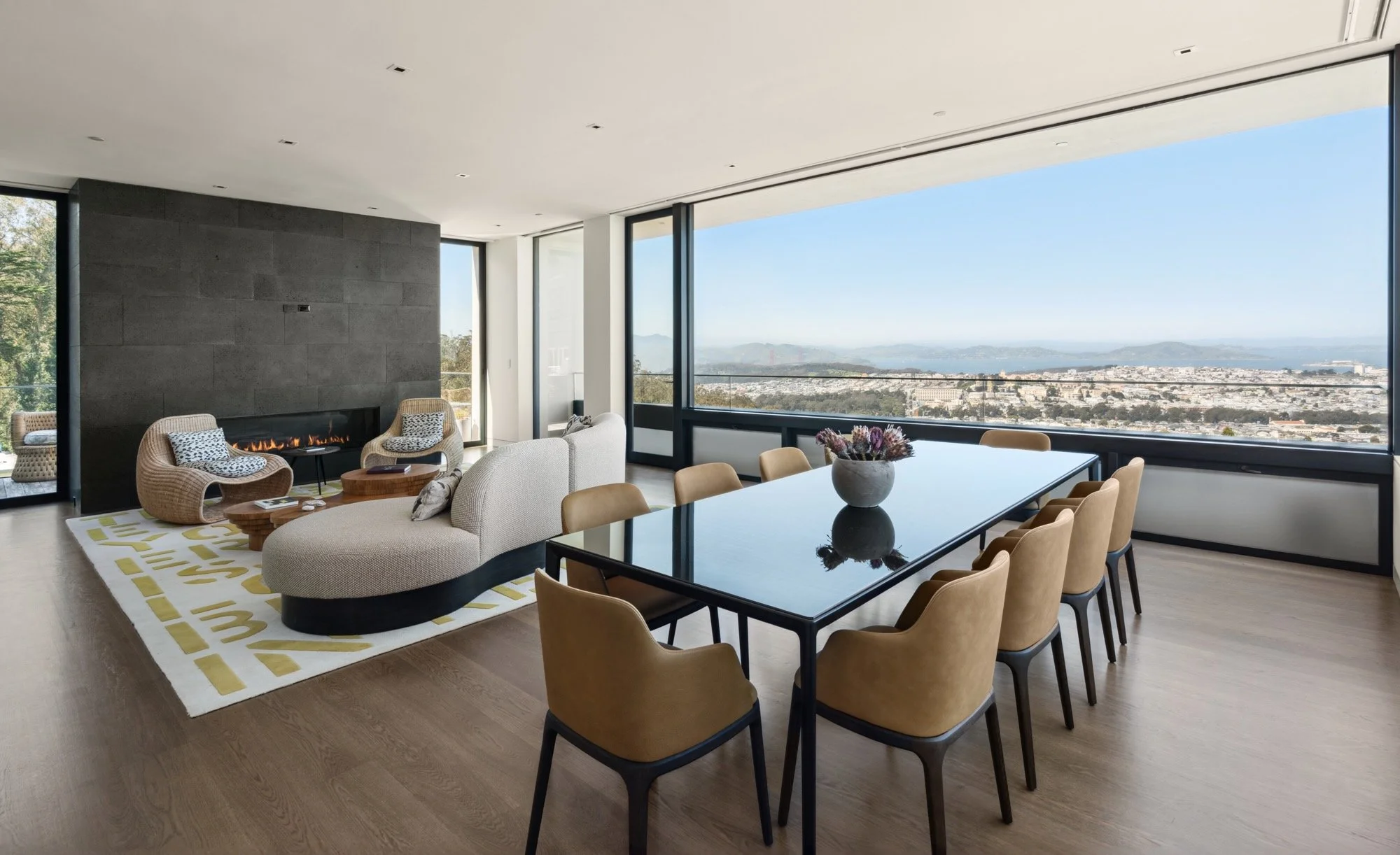 Modern living and dining room with large windows showing city and mountain views, a fireplace, cream sofa, wicker chairs, a black dining table with beige chairs, and decorative pillows and flowers.