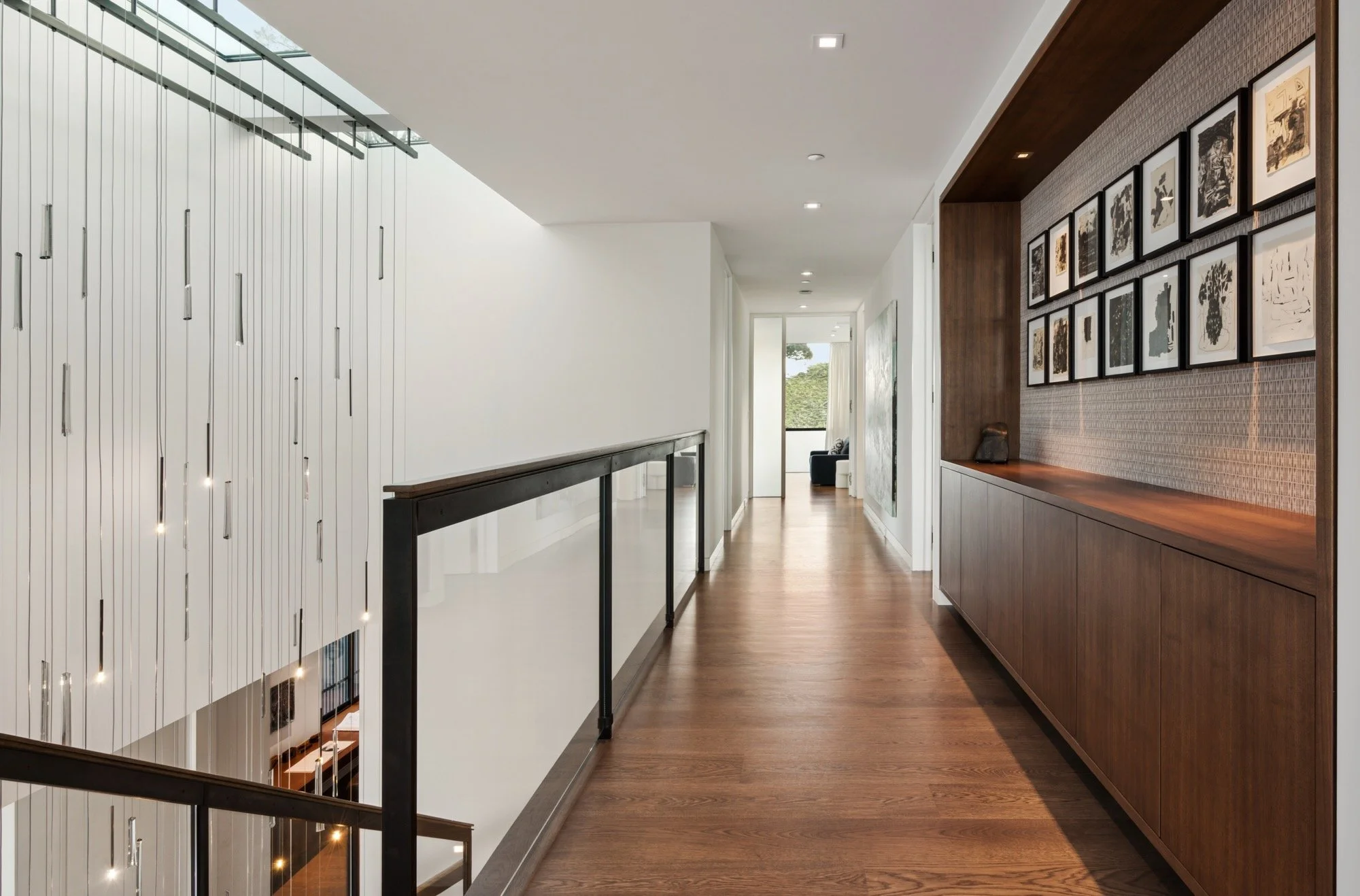 Interior view of a modern house hallway with wooden flooring, framed artwork on the wall, a railing on the left, and a glimpse of a seating area with a window in the background.
