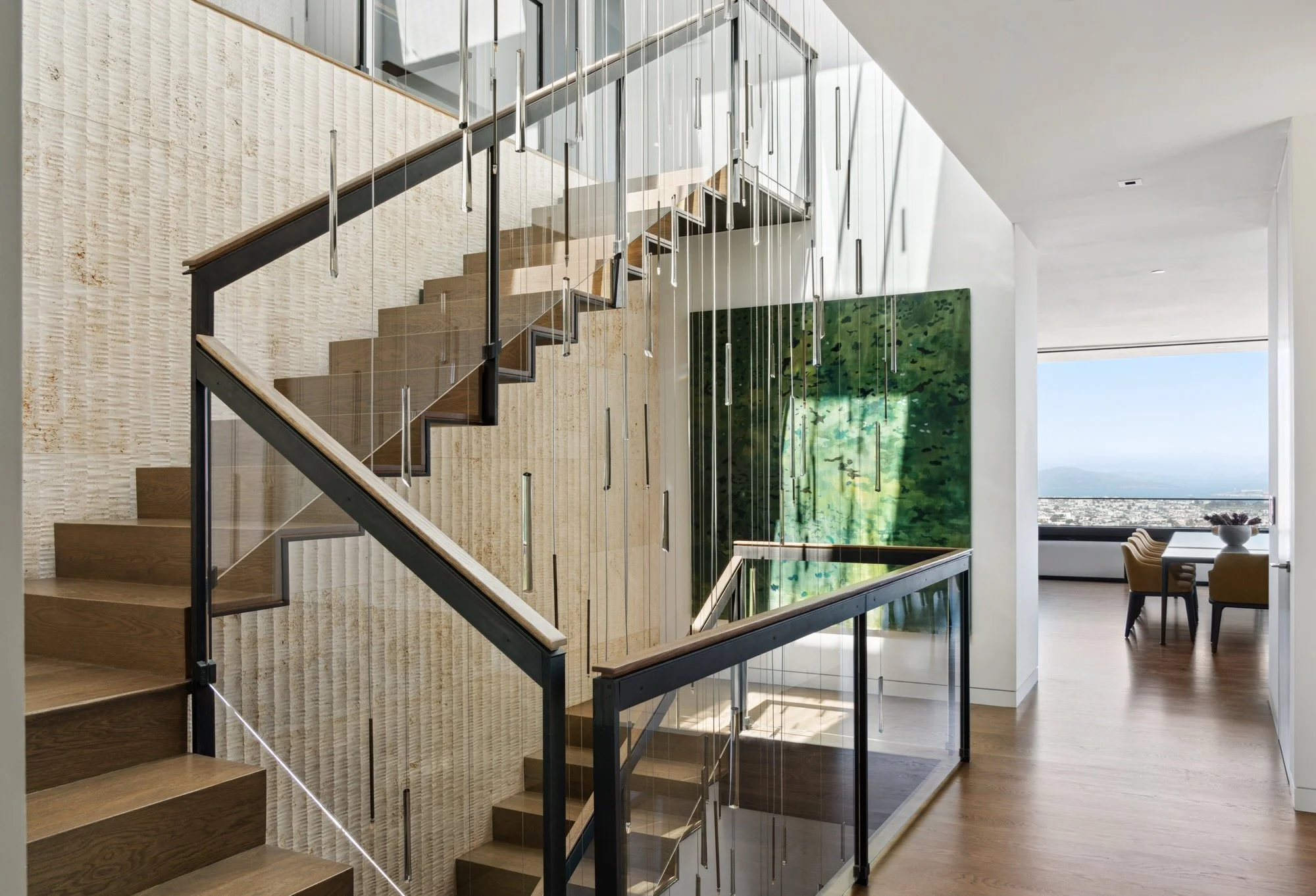 Modern staircase with glass railing and hanging decorative light fixtures in a spacious, sunlit interior with artwork and city view