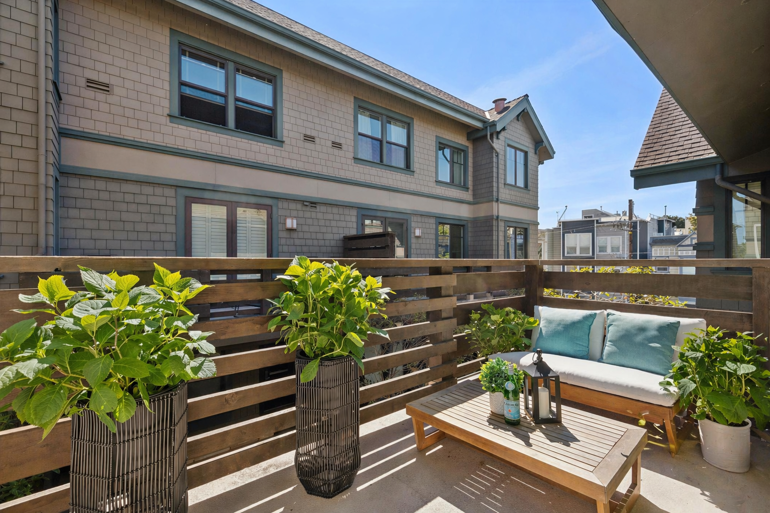 A cozy balcony with potted green plants, a wooden sofa with teal pillows, a wooden coffee table with lanterns and a bottle, surrounded by a wooden fence, overlooking neighboring rooftops and a blue sky.