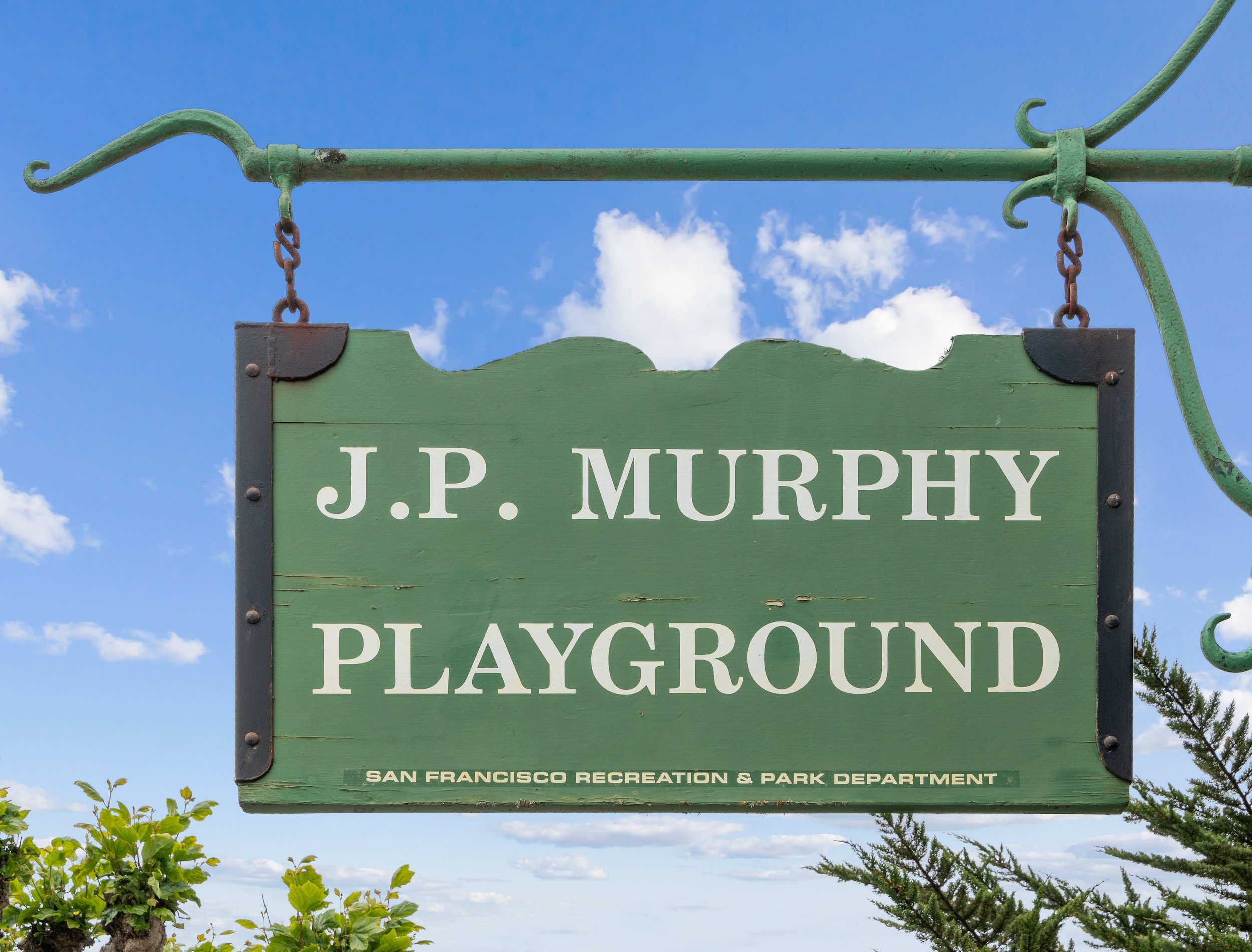 Green wooden sign hanging from a metal frame against a blue sky with clouds, reading "J.P. Murphy Playground" and indicating it is managed by the San Francisco Recreation & Park Department.
