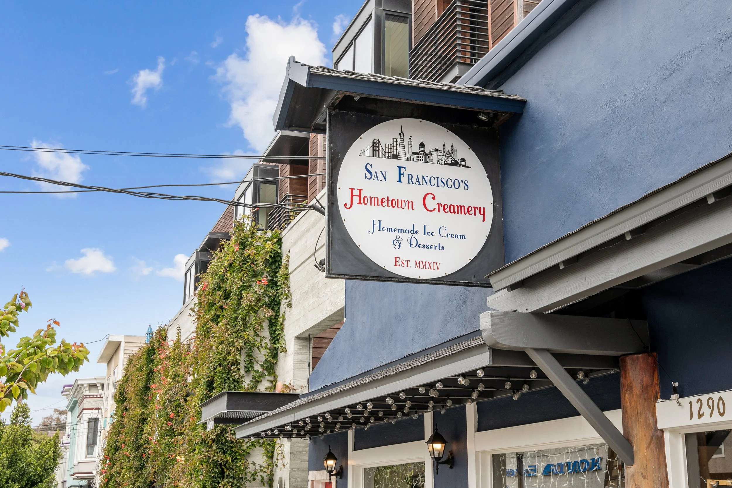 A street view of a building with a sign for San Francisco's Hometown Creamery, advertising homemade ice cream and desserts, established in 1914. The building features a shaded sidewalk with string lights, and there are flowering plants along the buil